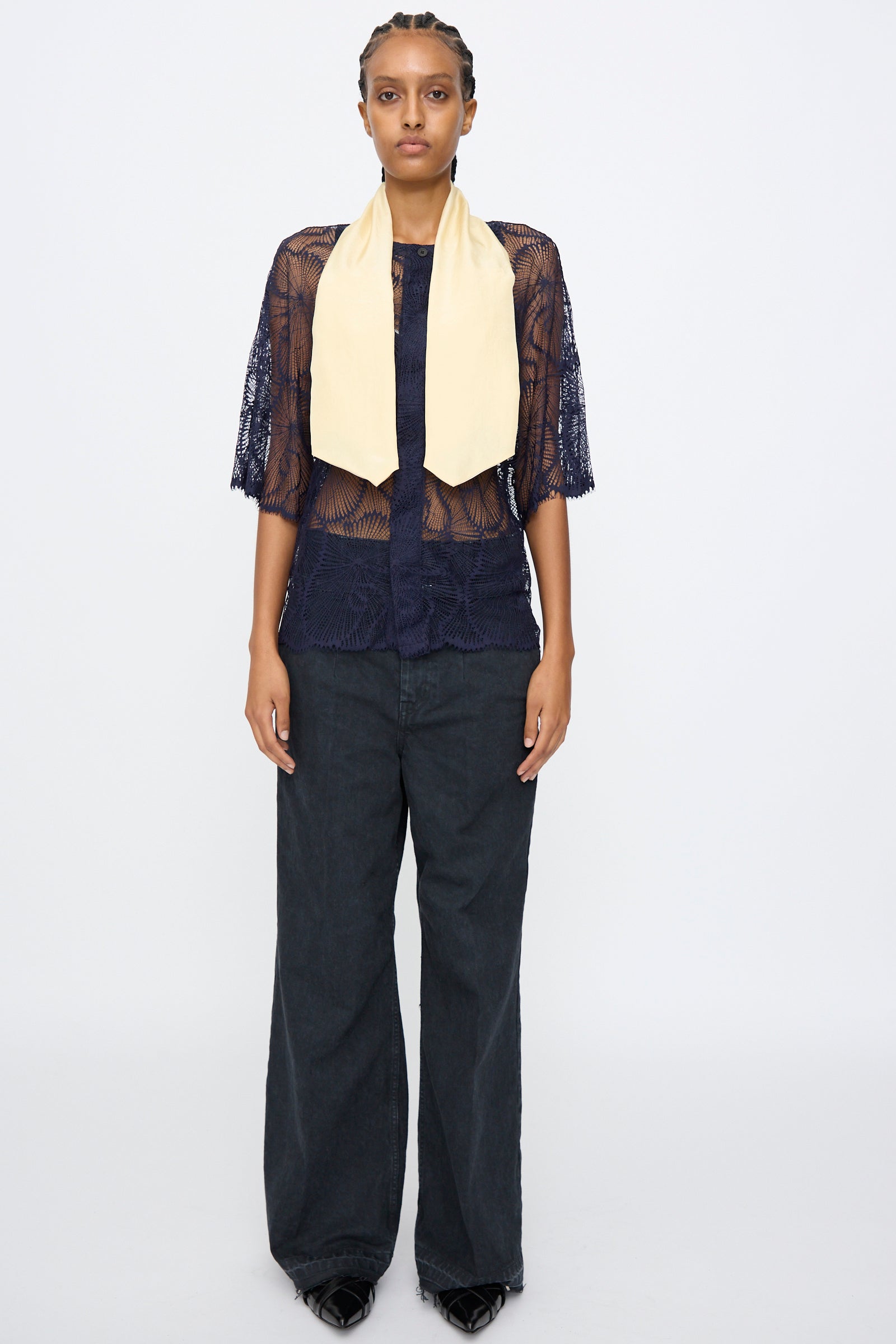 A person faces forward wearing TOGA ARCHIVES’ sheer navy Lace Blouse with an oversized fit, wide-leg black pants, black shoes, and a light yellow scarf draped around the neck.