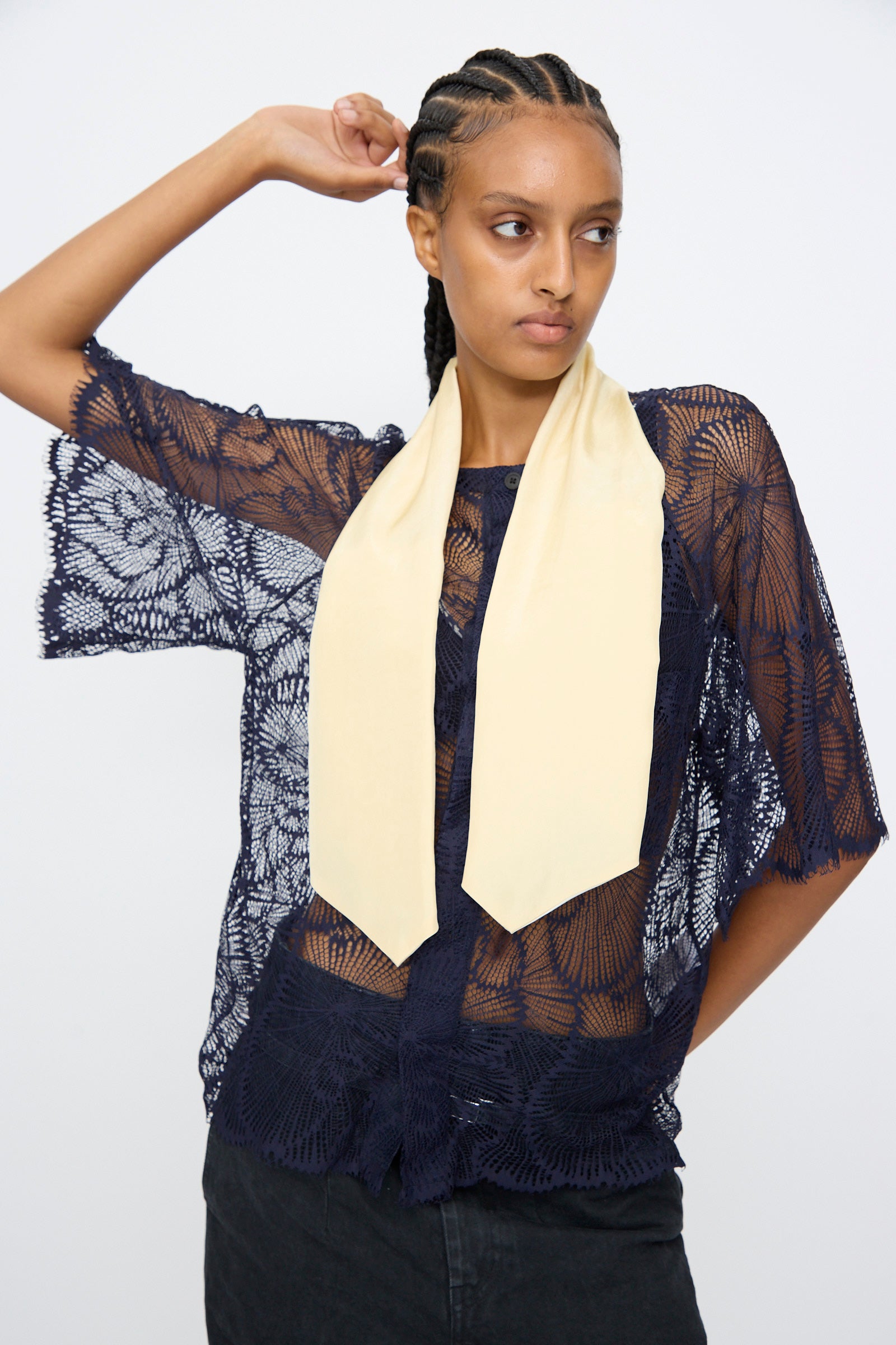 A model poses with one arm raised against a plain background, wearing the TOGA ARCHIVES Lace Blouse in Navy, styled with a cream scarf and black bottoms.