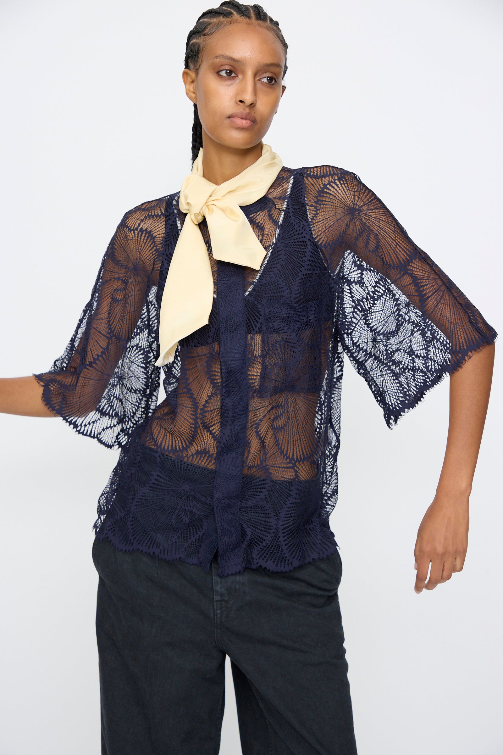 A person models the TOGA ARCHIVES Lace Blouse in Navy, styled in an oversized sheer fit over a dark top with black pants and a light yellow neck scarf, standing against a plain background.