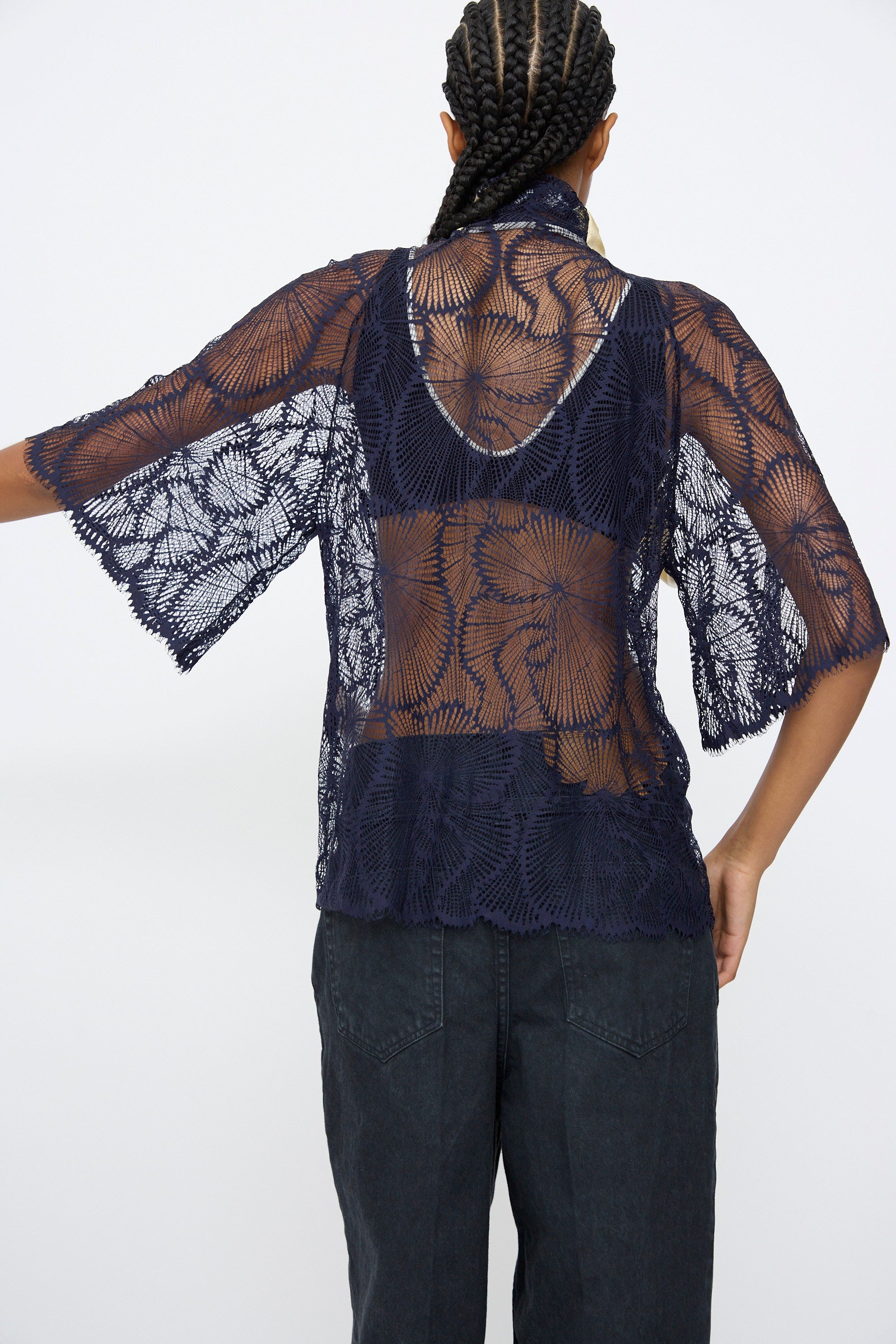 Person faces away, wearing the TOGA ARCHIVES Lace Blouse in Navy—an oversized sheer lace top over a black undergarment and black pants, with braided hair.