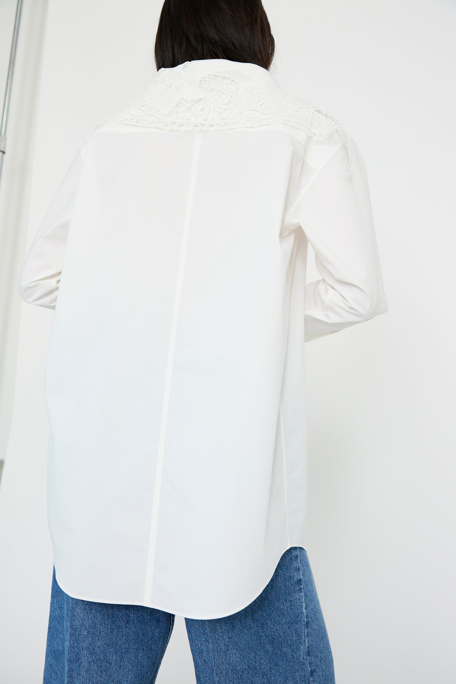 A person in a TOGA ARCHIVES Lace Boat Neck Shirt in White and blue jeans is seen from the back against a plain backdrop.