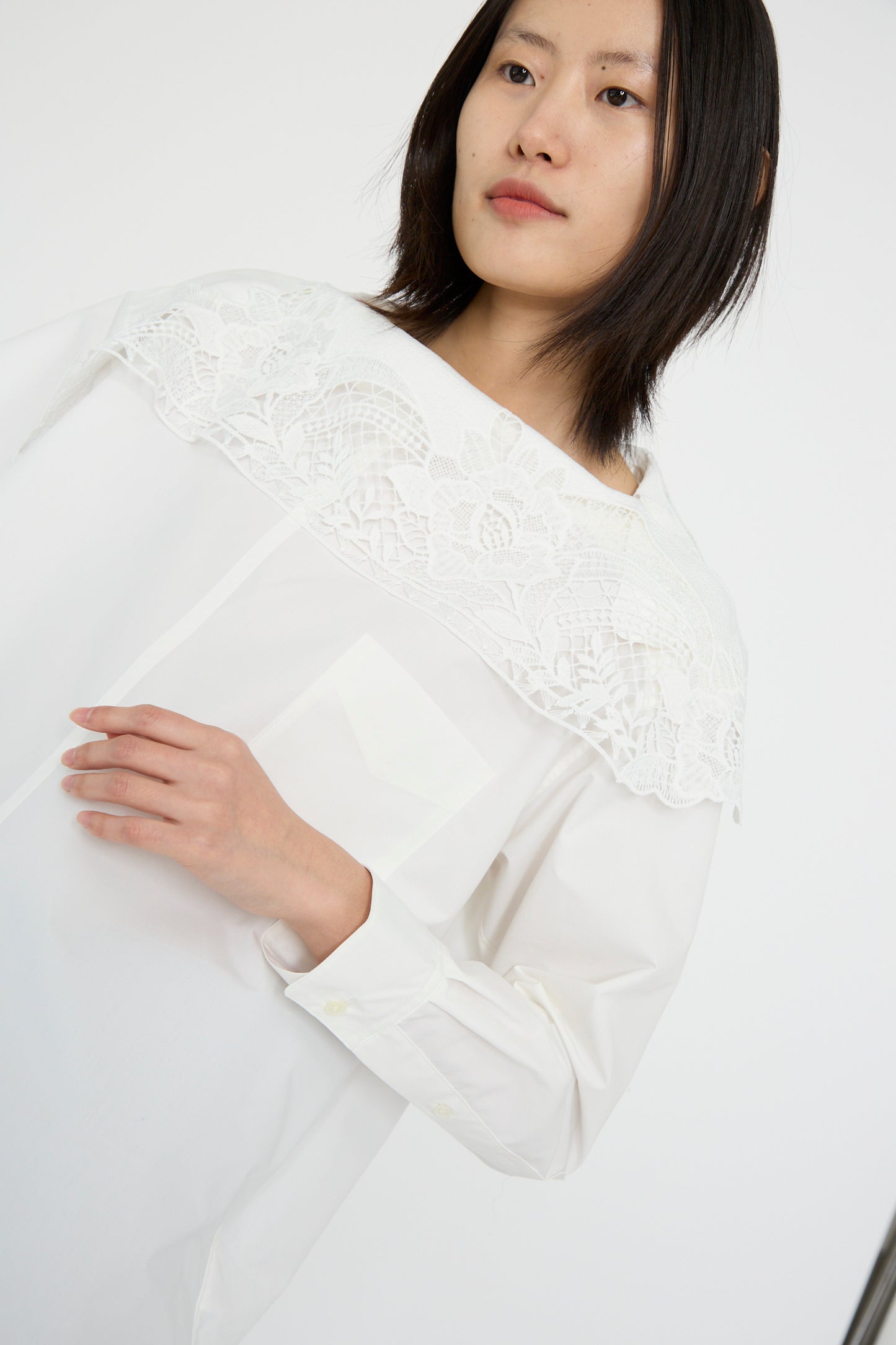 A person in a TOGA ARCHIVES Lace Boat Neck Shirt in white, with an oversized fit with a delicate lace collar, stands against a simple background.