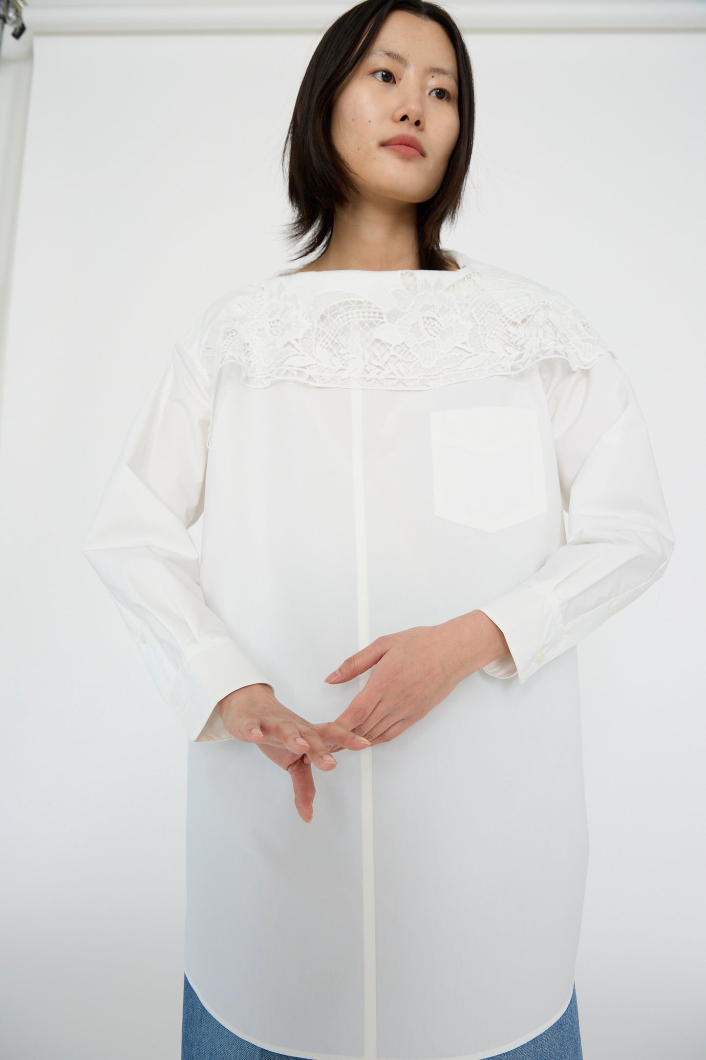 A person wearing a Lace Boat Neck Shirt in White by TOGA ARCHIVES and blue jeans stands against a plain background, hands clasped.