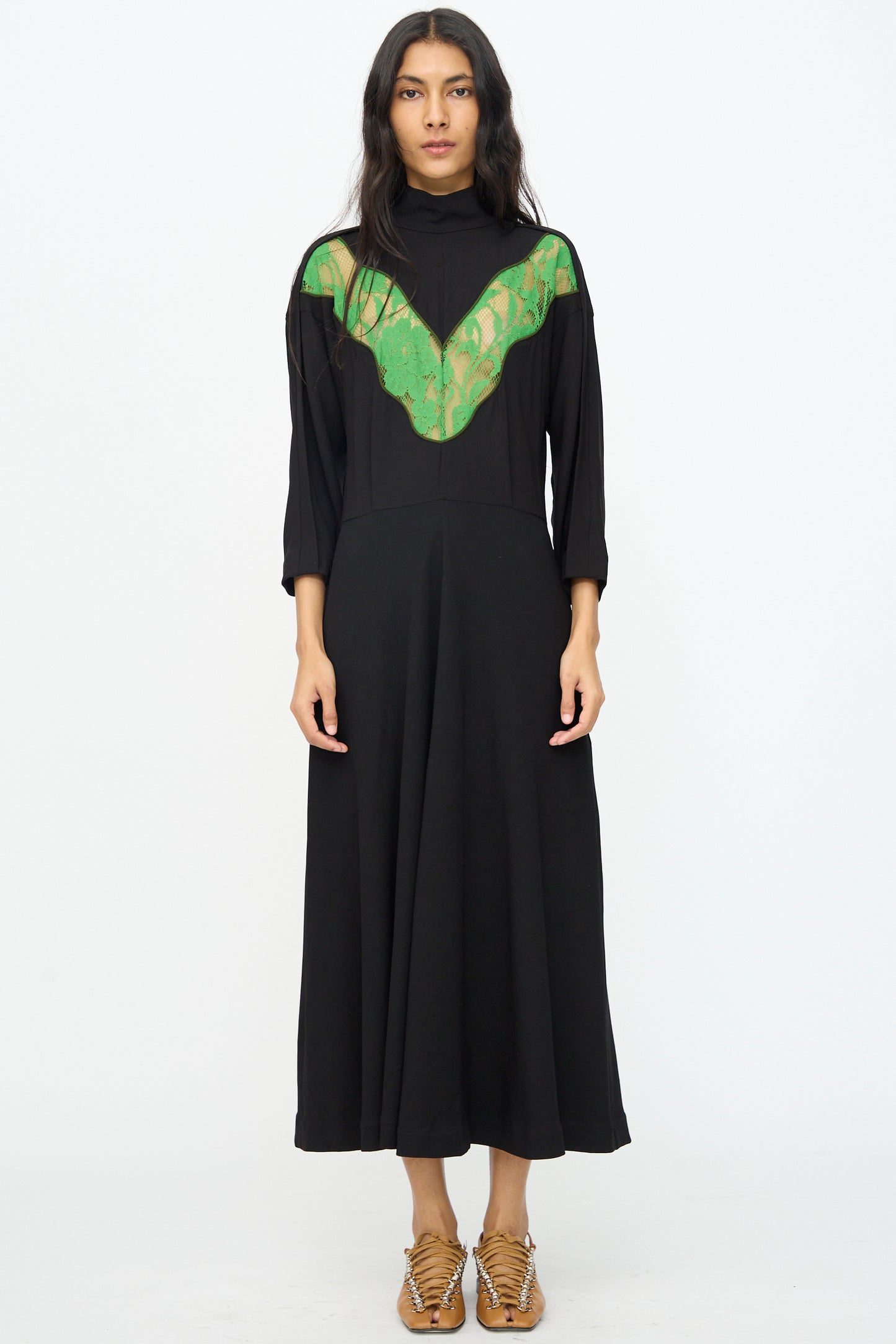 A woman stands against a white background, wearing the TOGA ARCHIVES Lace Dress in Black with long sleeves and lace detail, paired with tan woven sandals.