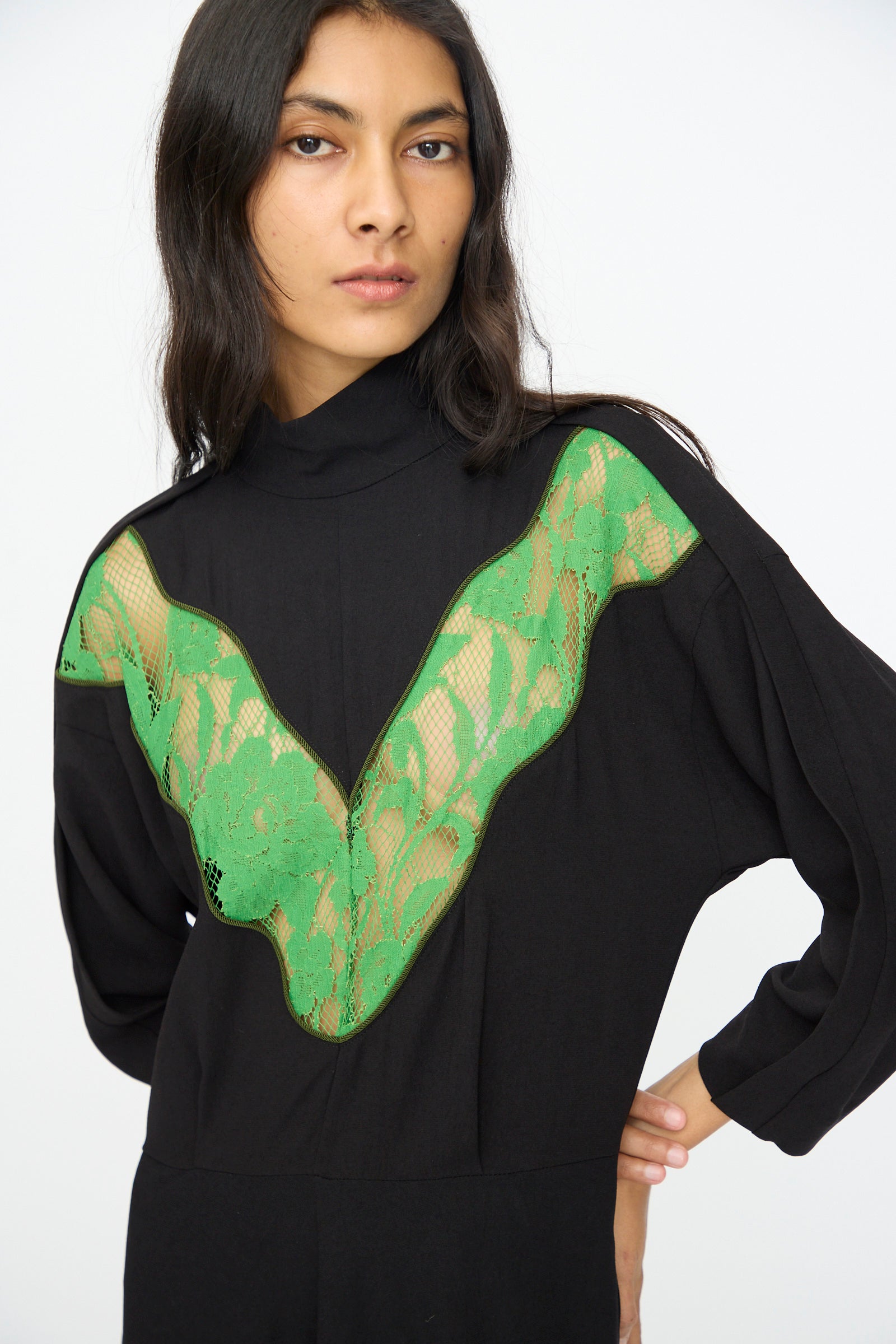 A woman with long dark hair wears TOGA ARCHIVES' Lace Dress in Black, an acetate blend design with a green lace V-shaped chest panel, standing against a plain light background.