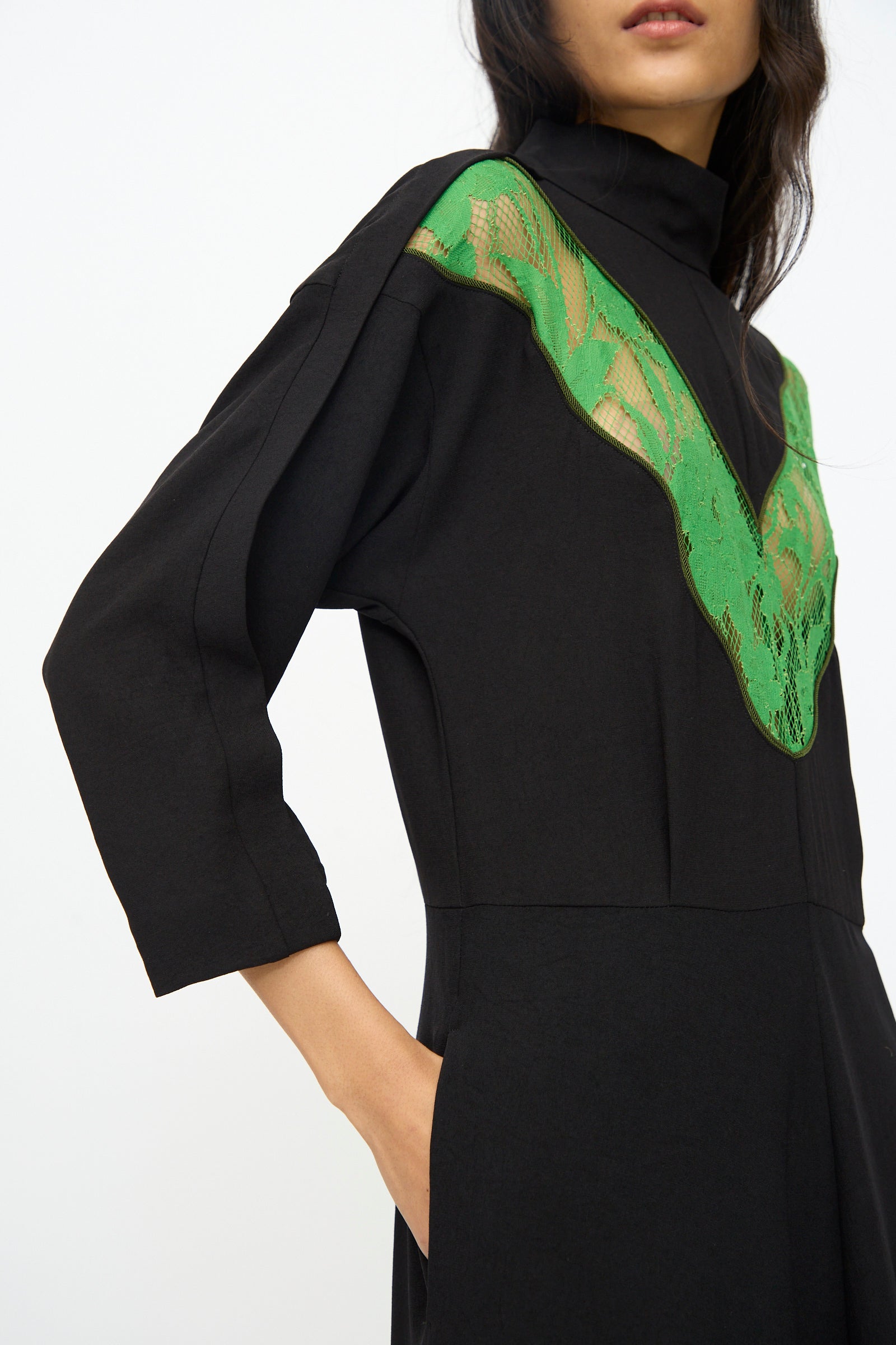 A woman wears the TOGA ARCHIVES Lace Dress in Black, a long sleeve midi style featuring green lace detail across the upper chest and shoulders, with one hand in her pocket.