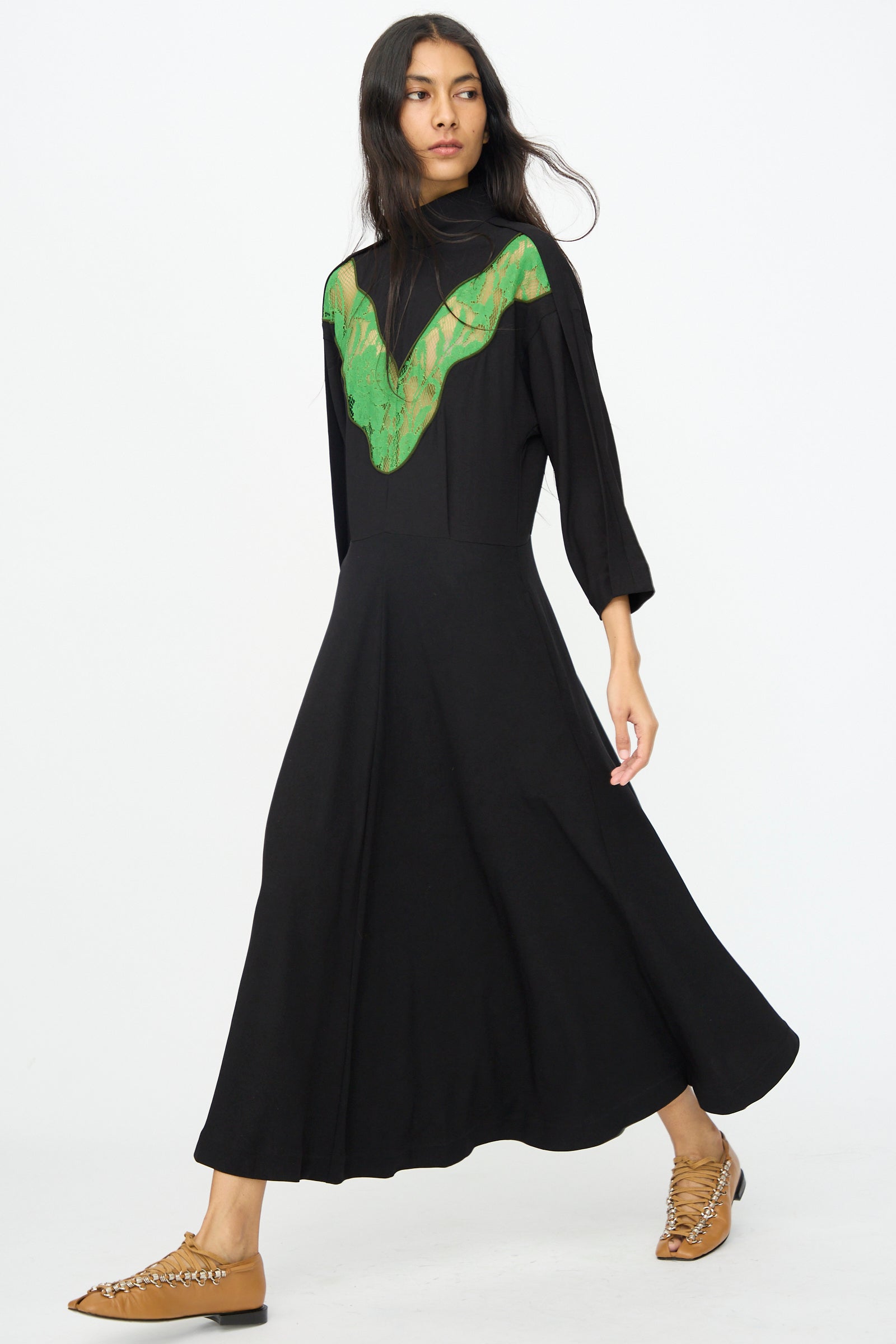 A woman wearing the TOGA ARCHIVES Lace Dress in Black with long sleeves, walking forward in tan flat shoes against a plain white background.