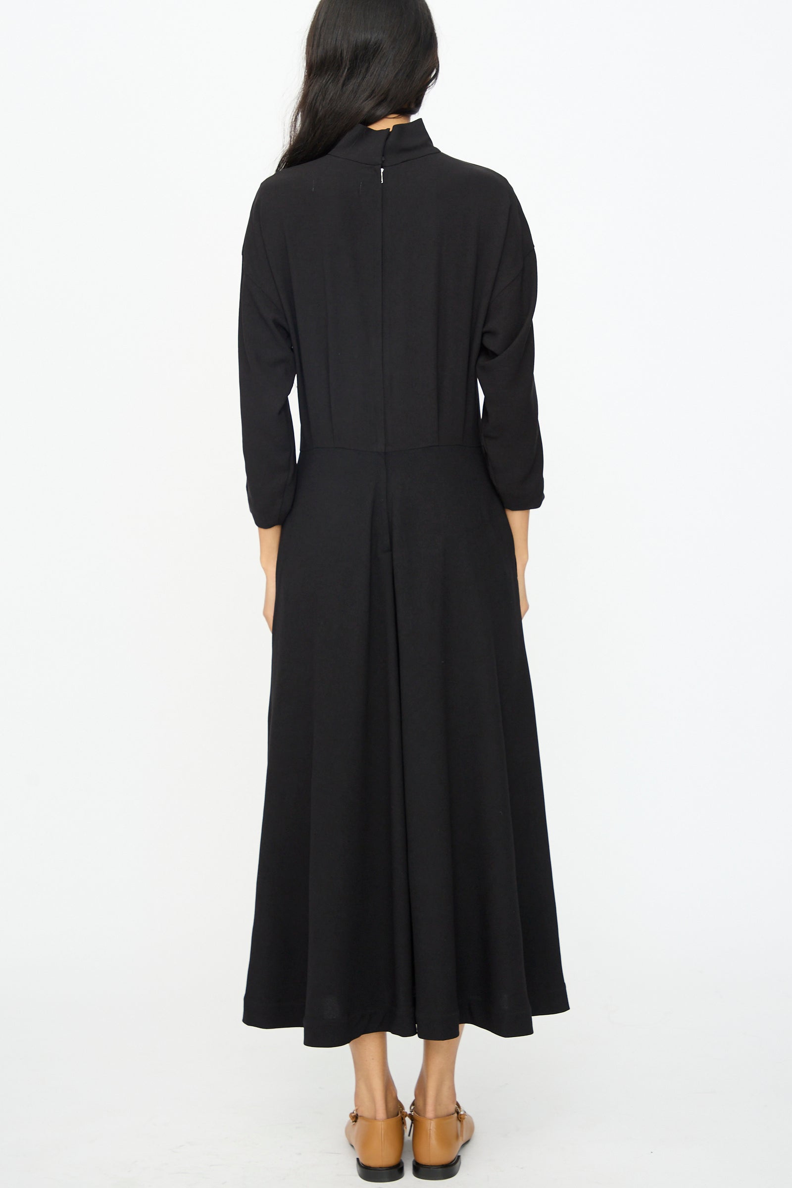 A woman with long dark hair stands with her back to the camera against a white background, wearing the TOGA ARCHIVES Lace Dress in Black and brown flats.