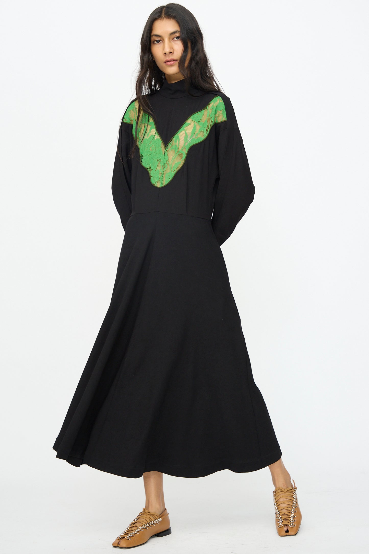 A woman wears the TOGA ARCHIVES Lace Dress in Black with a green lace V-shaped chest detail and tan lace-up shoes, standing against a plain white background.