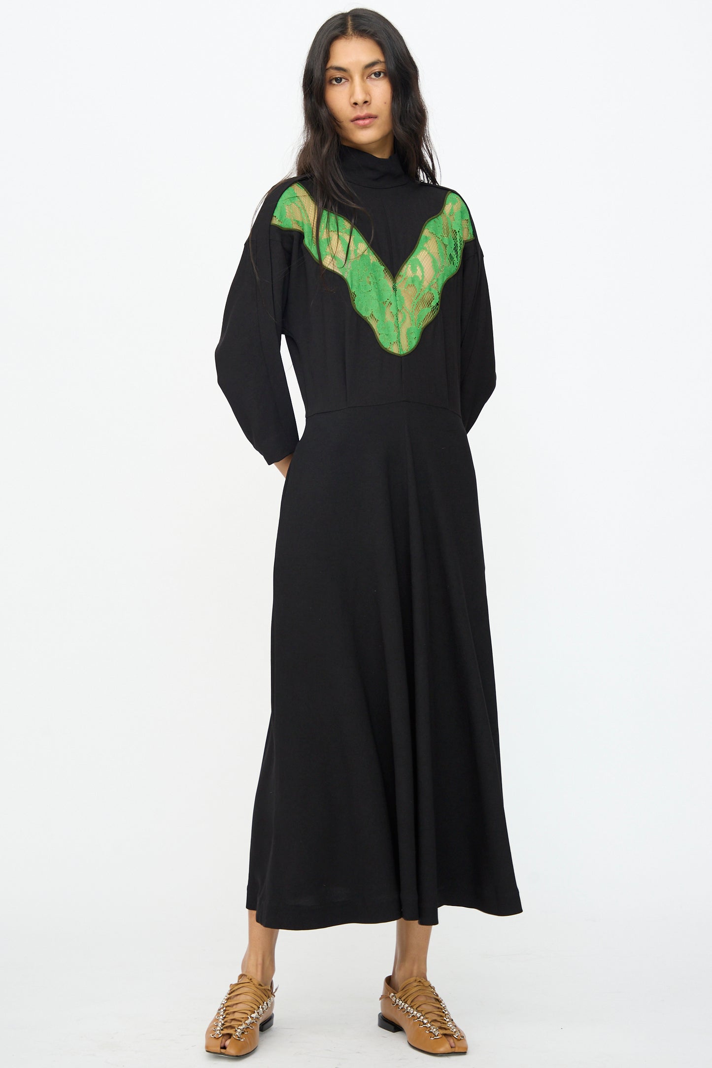 A woman stands in the Lace Dress in Black by TOGA ARCHIVES, featuring long sleeves and lace detail on the chest, paired with tan woven shoes against a plain white background.