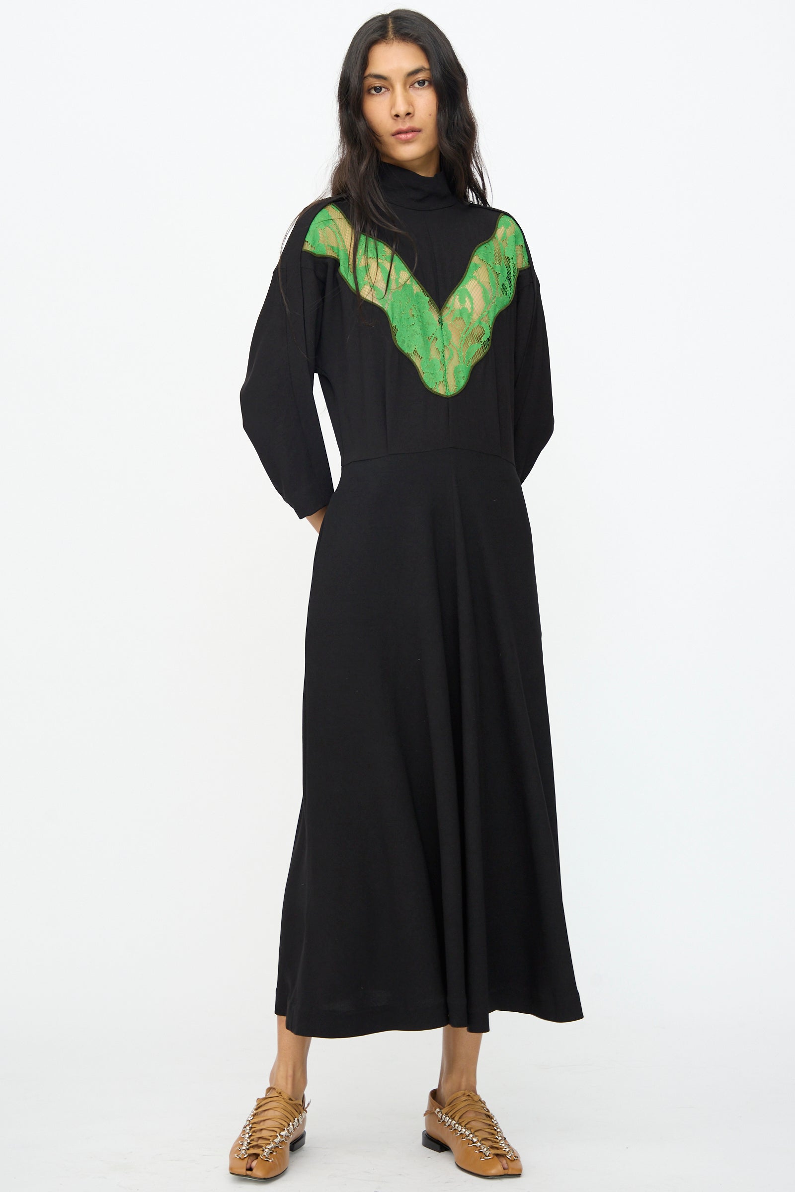 A woman stands in the Lace Dress in Black by TOGA ARCHIVES, featuring long sleeves and lace detail on the chest, paired with tan woven shoes against a plain white background.