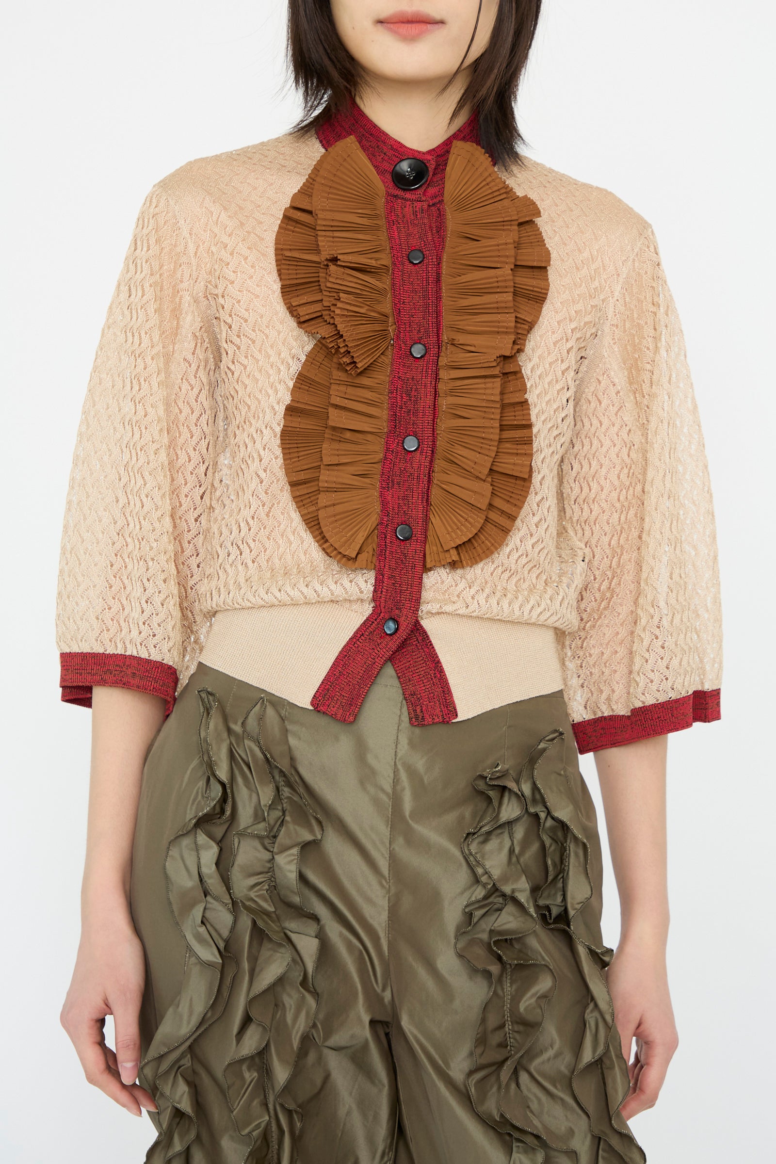 A model is wearing a Lace Knit Cardigan in Beige by TOGA ARCHIVES, featuring textured ruffled brown and red accents, paired with olive green pants with ruffled details, facing forward against a white backdrop. 