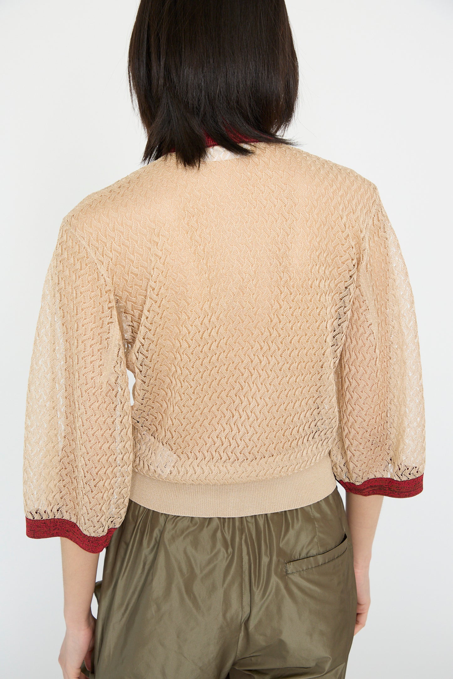 A person with dark shoulder-length hair is seen from behind, wearing a sheer beige top with red trim, high-waisted olive pants, and a TOGA ARCHIVES Lace Knit Cardigan in Beige.