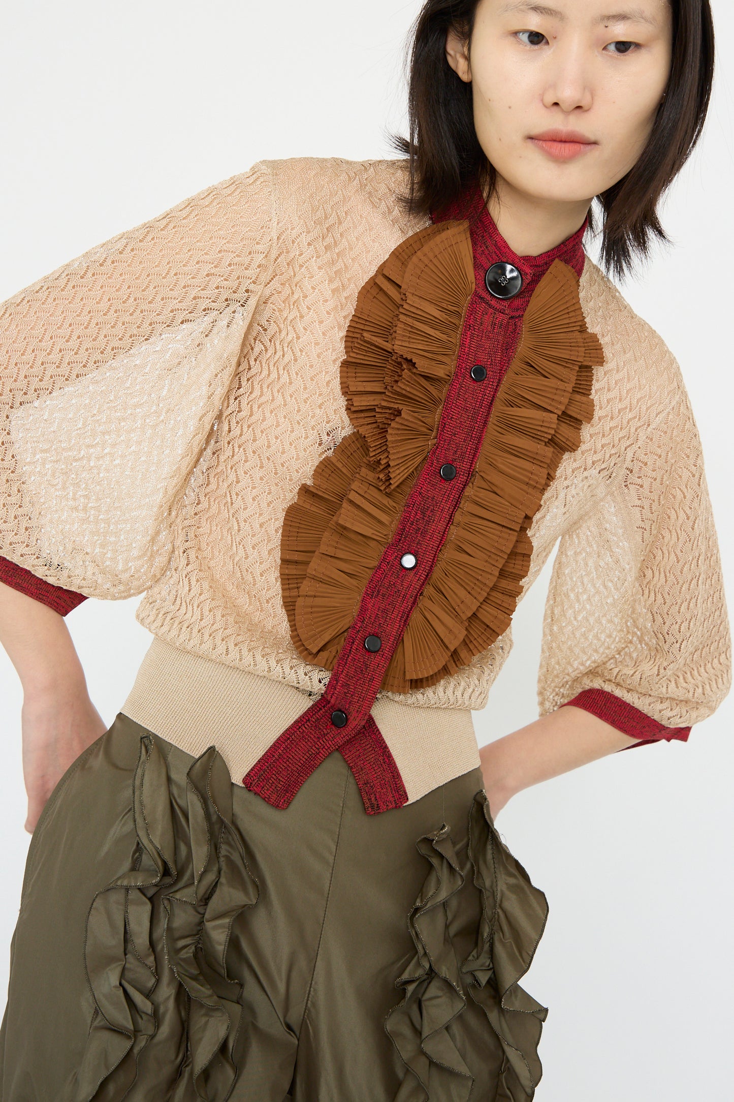 A woman is wearing the TOGA ARCHIVES Lace Knit Cardigan in Beige with brown ruffles and a high neck, paired with an olive skirt, against a plain background.