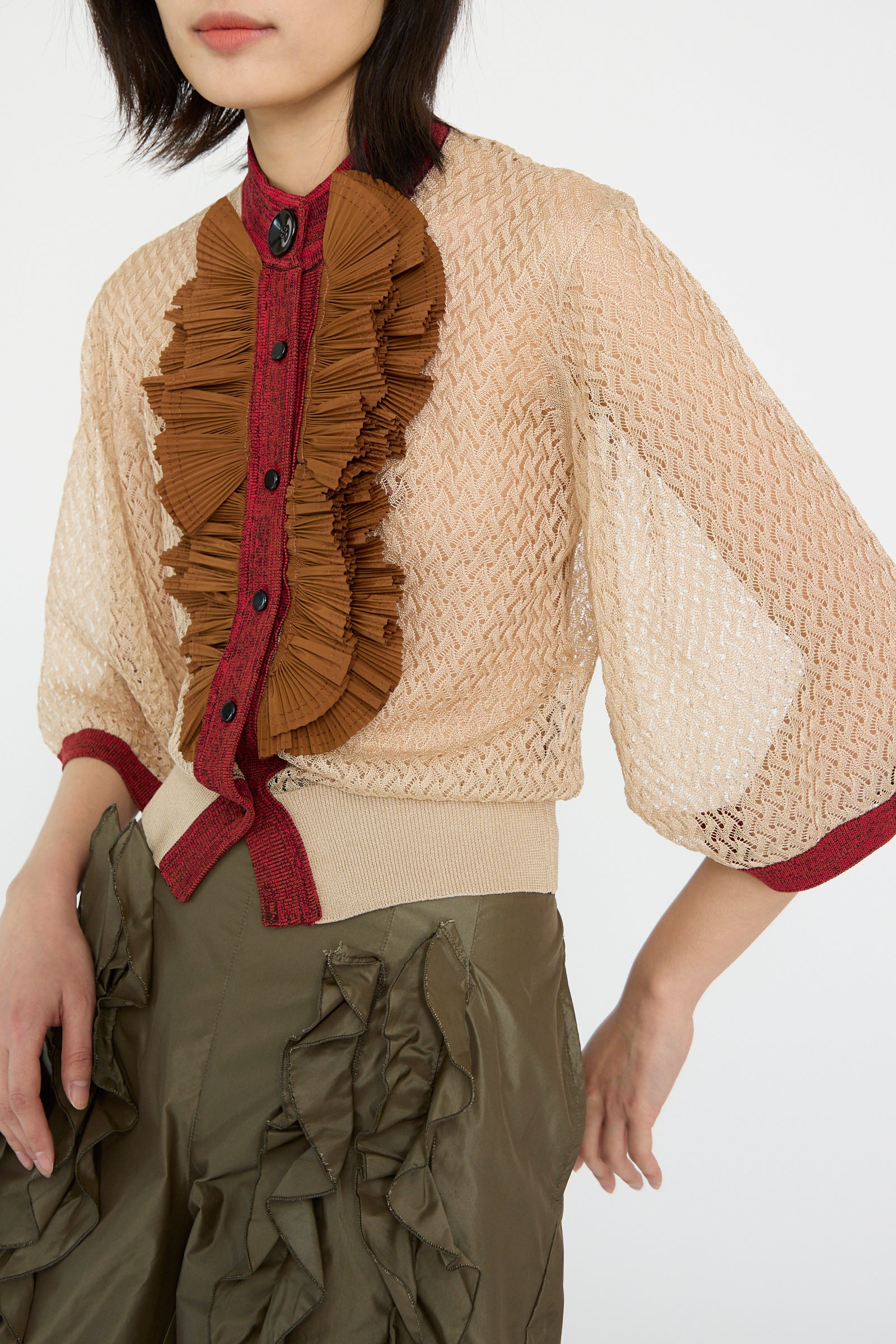 A person is wearing a beige Lace Knit Cardigan by TOGA ARCHIVES over a textured top with brown ruffles and dark red accents, paired with olive green pants featuring multiple ruffles.