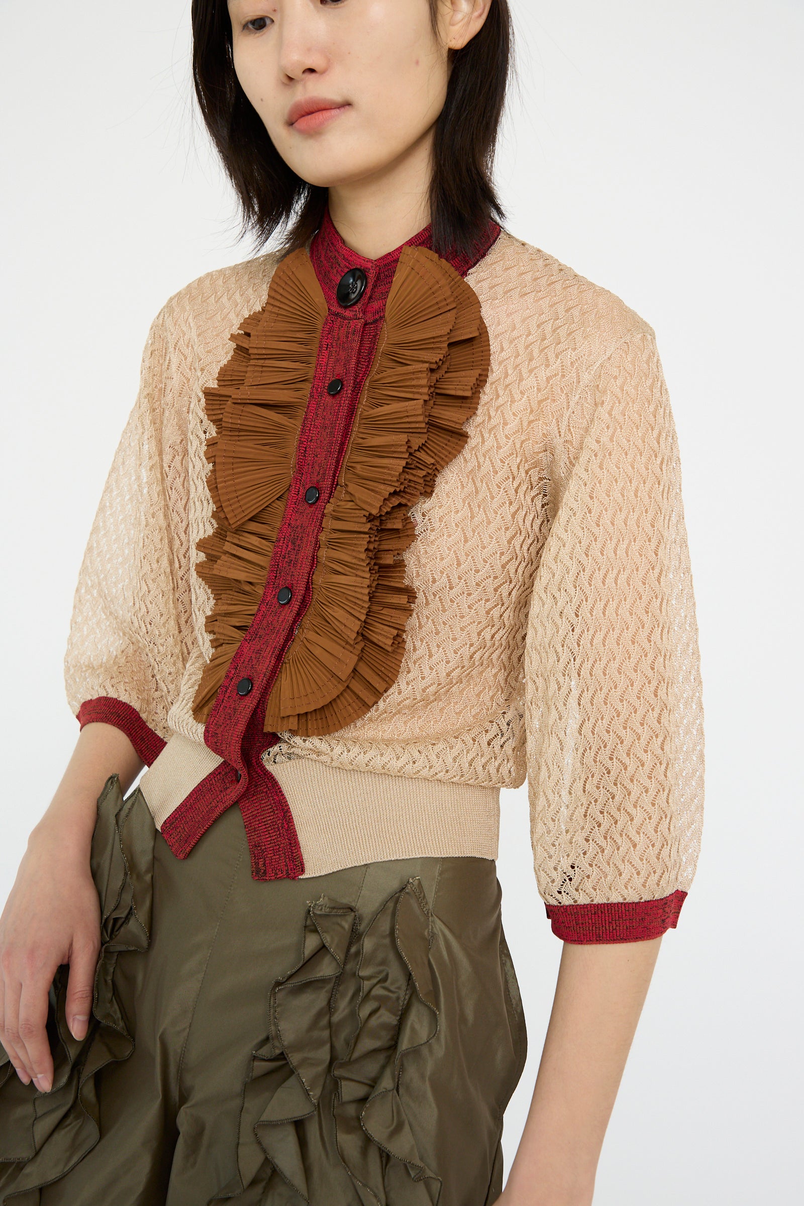 A person is wearing the TOGA ARCHIVES Lace Knit Cardigan in beige, featuring a 3/4 sleeve design with a high neck and red and brown ruffled front, paired with olive green pants that also have ruffles.