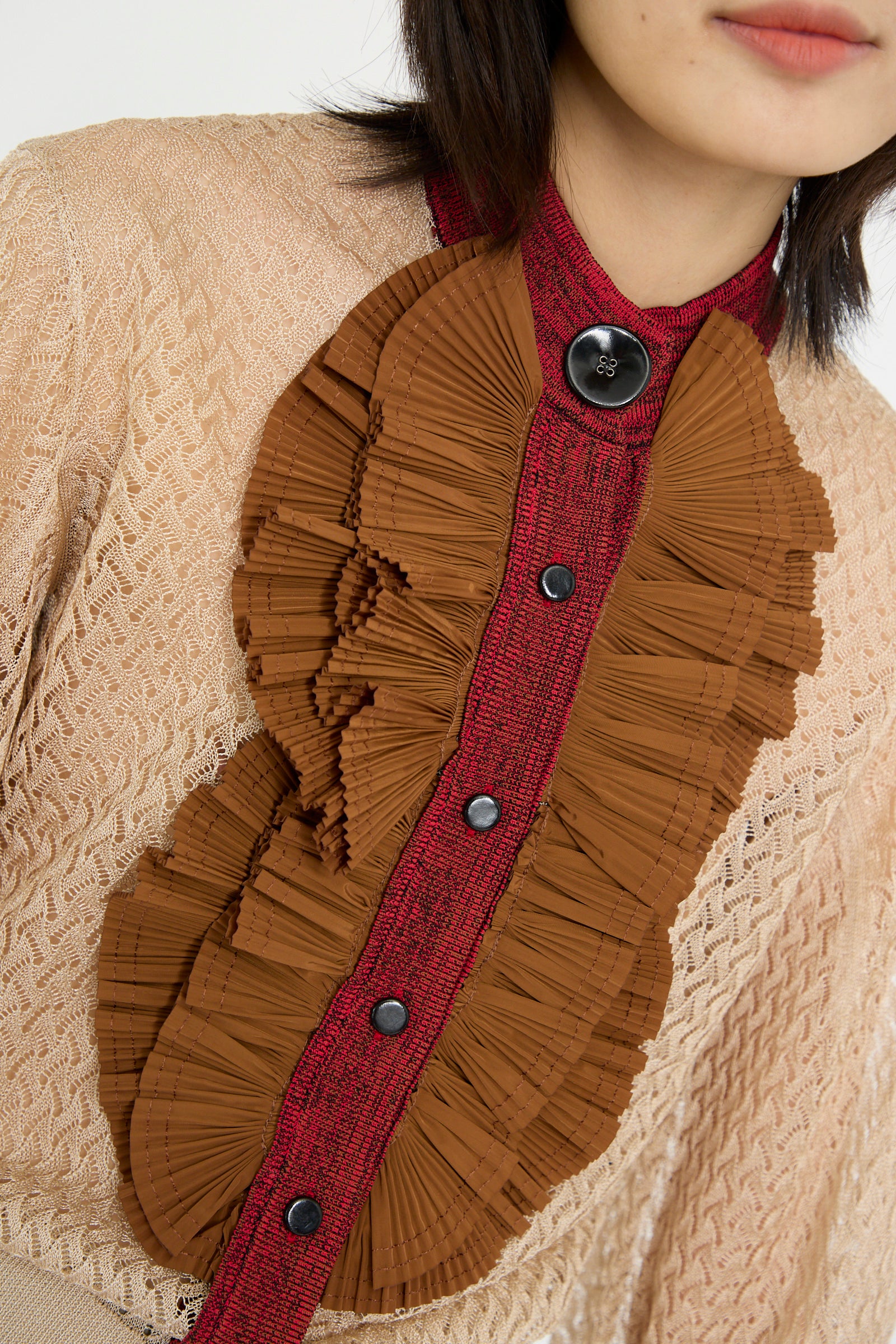 Close-up of a person wearing the TOGA ARCHIVES Lace Knit Cardigan in Beige, featuring a high neck, red button-down center, and brown pleated ruffles.