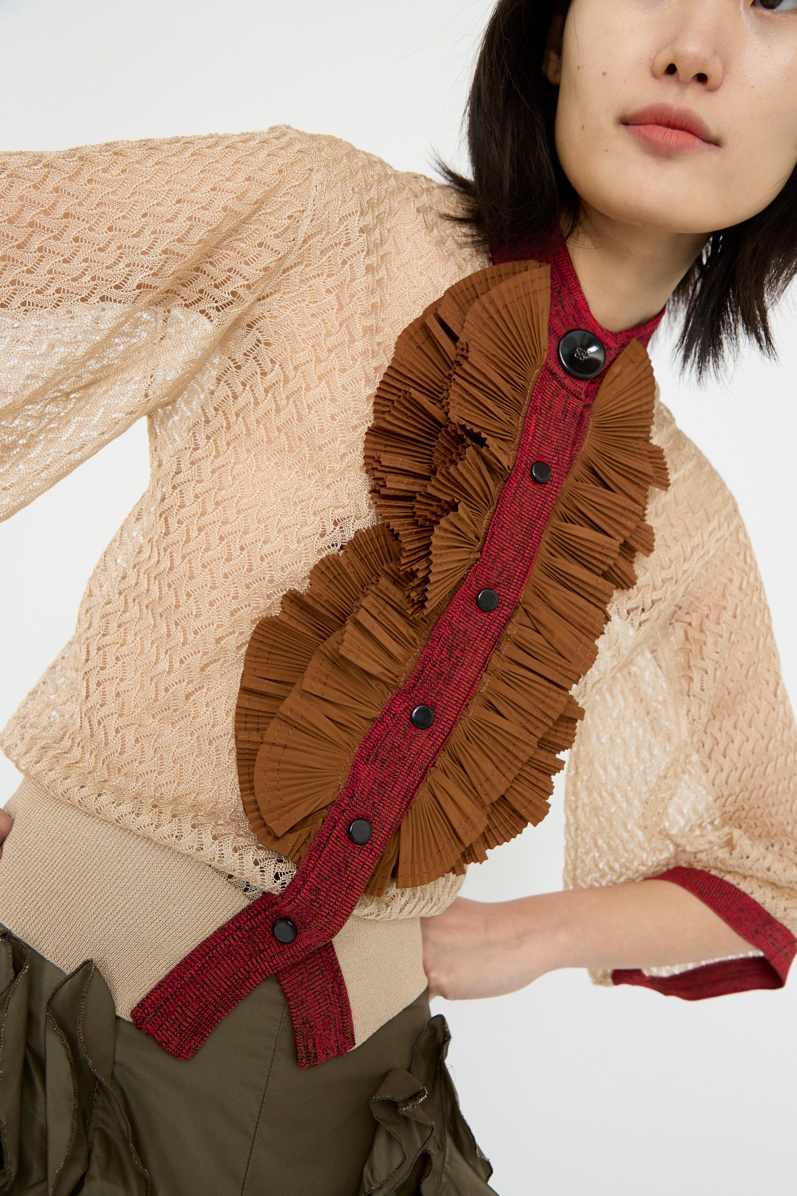 A model in a TOGA ARCHIVES Lace Knit Cardigan in beige featuring brown ruffles and red trim, paired with olive green pants, against a white backdrop.