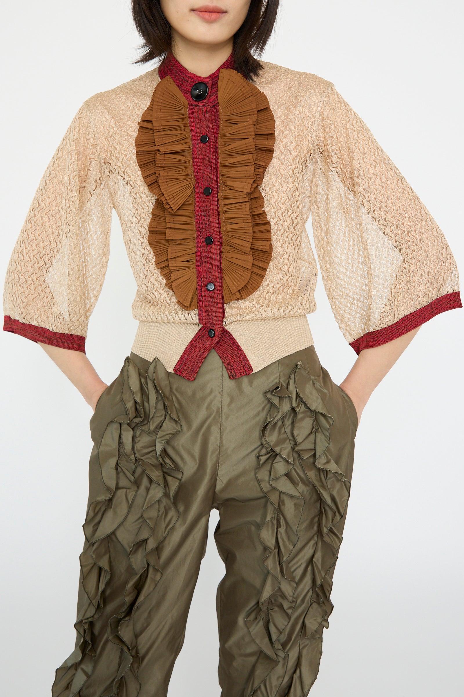 A person in a sheer, beige blouse with brown ruffles and olive green, ruffle-detailed pants stands against a plain background, wearing TOGA ARCHIVES Lace Knit Cardigan in Beige.