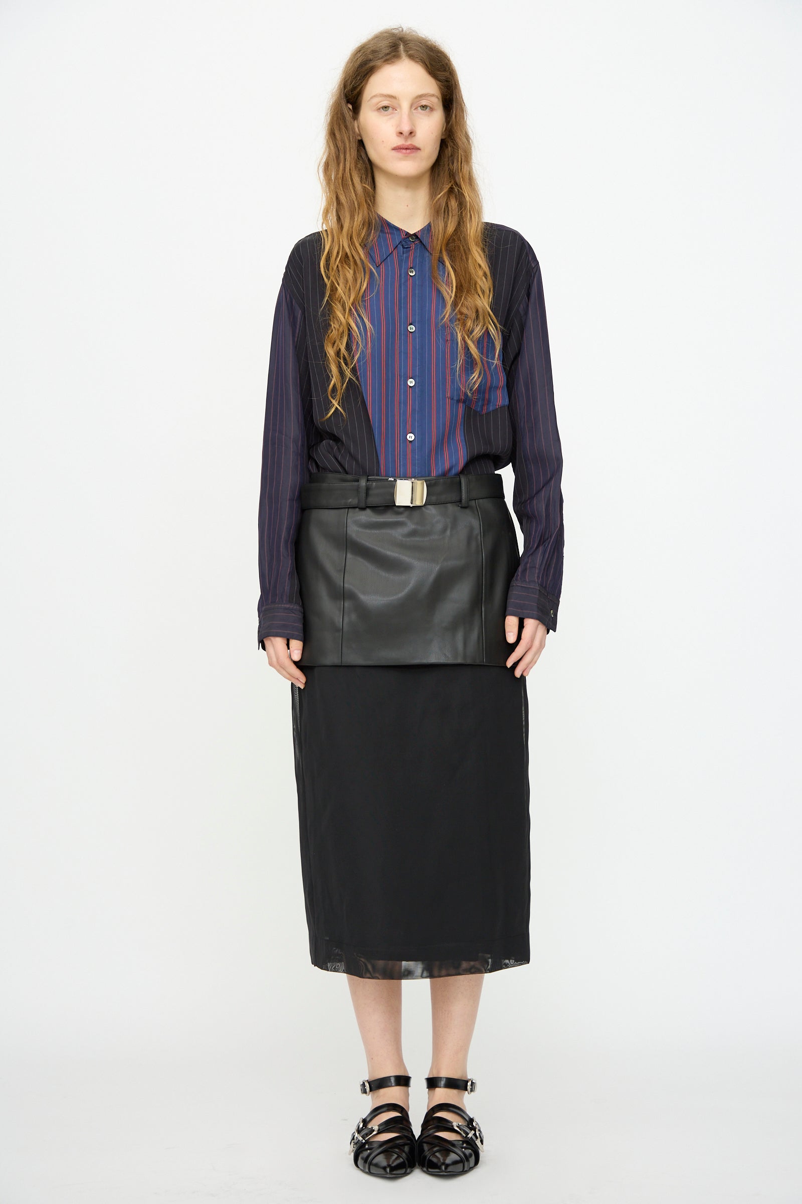 A person stands against a white background, wearing a striped button-up shirt, black sandals, and the TOGA ARCHIVES Leather Mesh Skirt in Black.