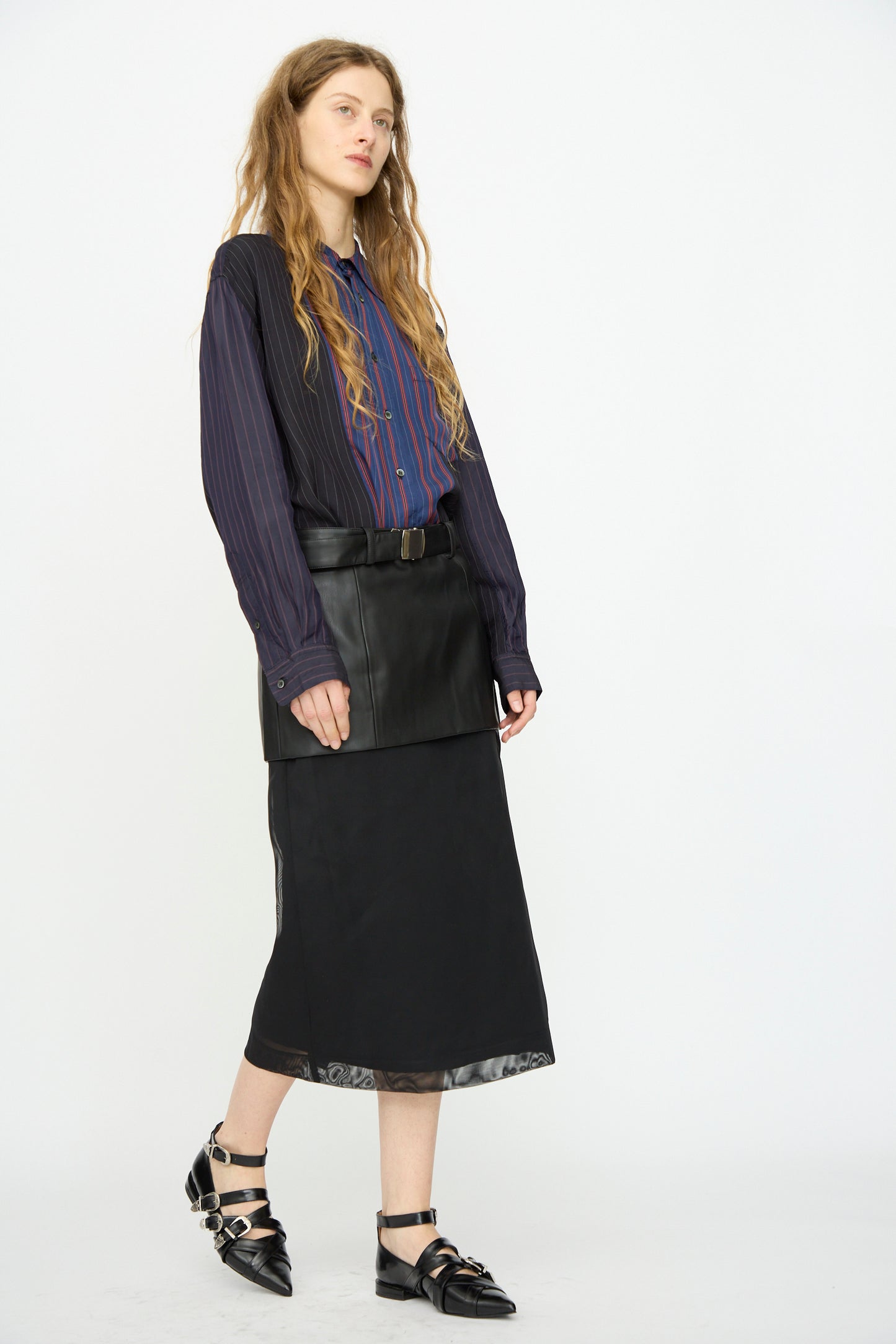 A person stands against a white background, wearing a striped button-up shirt, the TOGA ARCHIVES Leather Mesh Skirt in Black with belt detail, and pointed black buckle shoes.