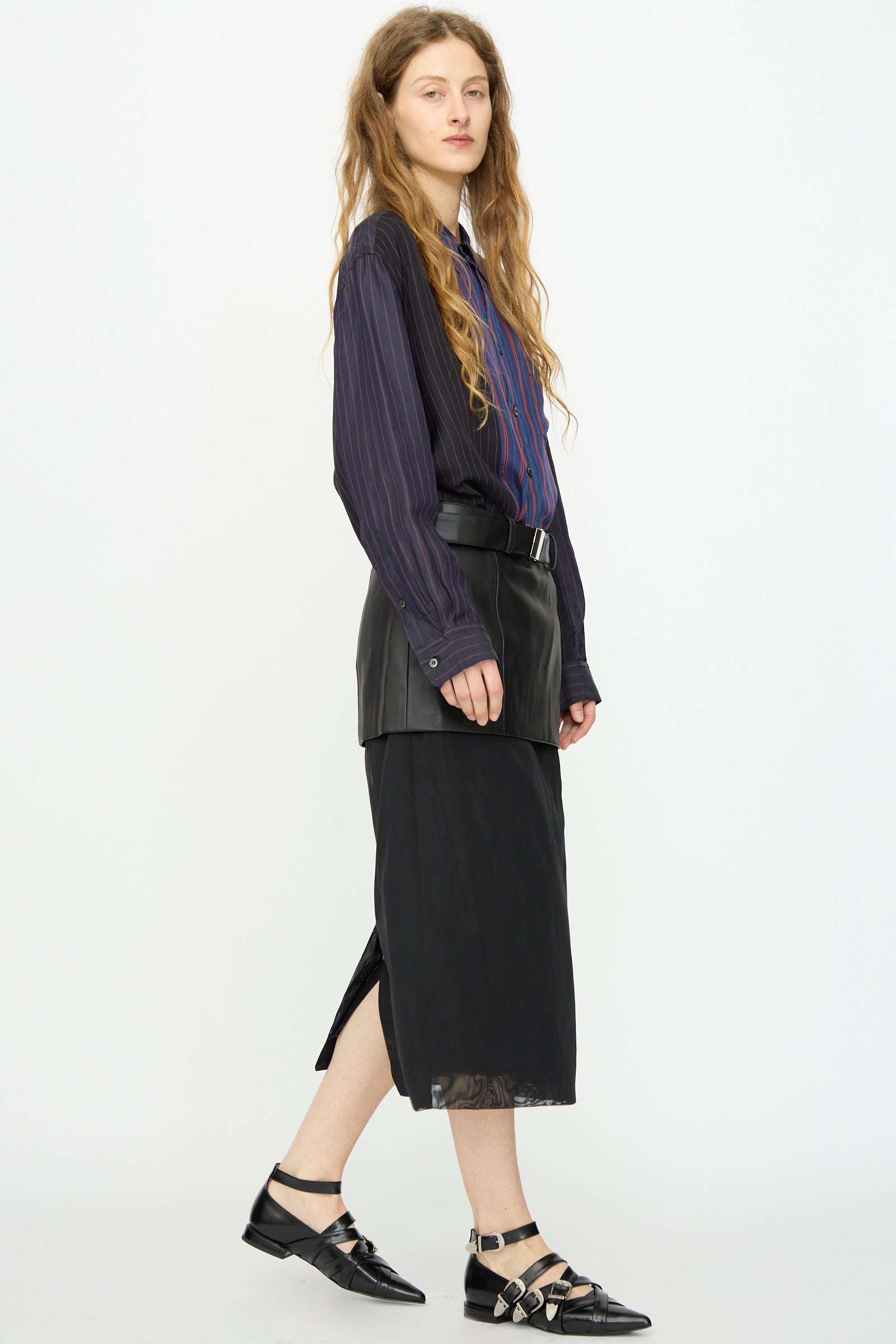 A person stands sideways against a white background, wearing a dark striped shirt, TOGA ARCHIVES Leather Mesh Skirt in Black, and black pointed shoes with buckles.