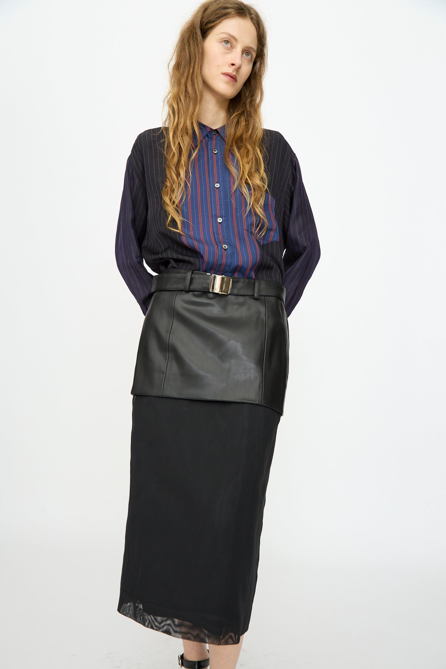 A person stands facing forward against a plain white background, wearing a blue and red striped shirt paired with the TOGA ARCHIVES Leather Mesh Skirt in Black.