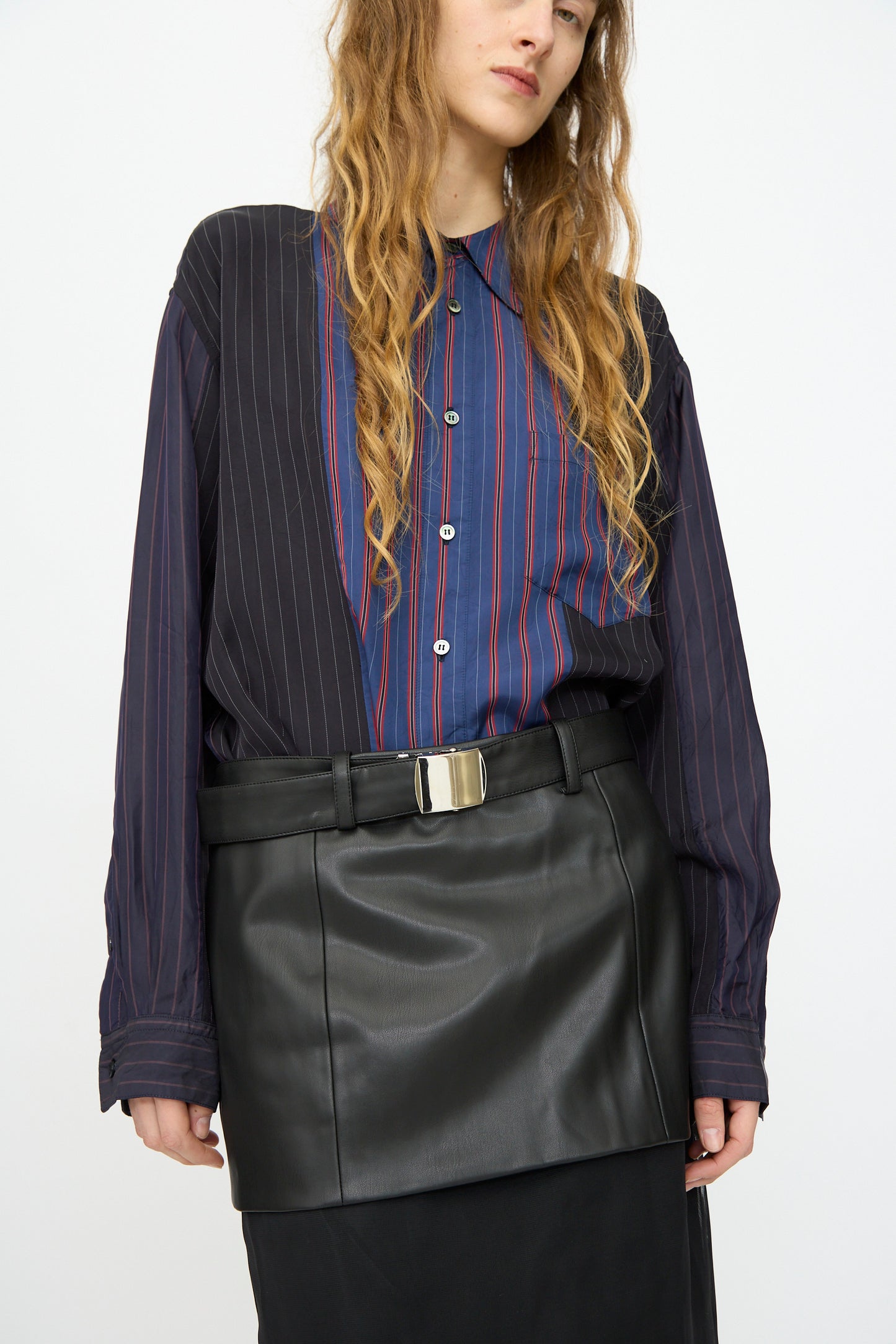 Person wears a blue and black pinstriped button-up shirt paired with the TOGA ARCHIVES Leather Mesh Skirt in Black, featuring a wide belt and silver buckle, standing against a plain background.