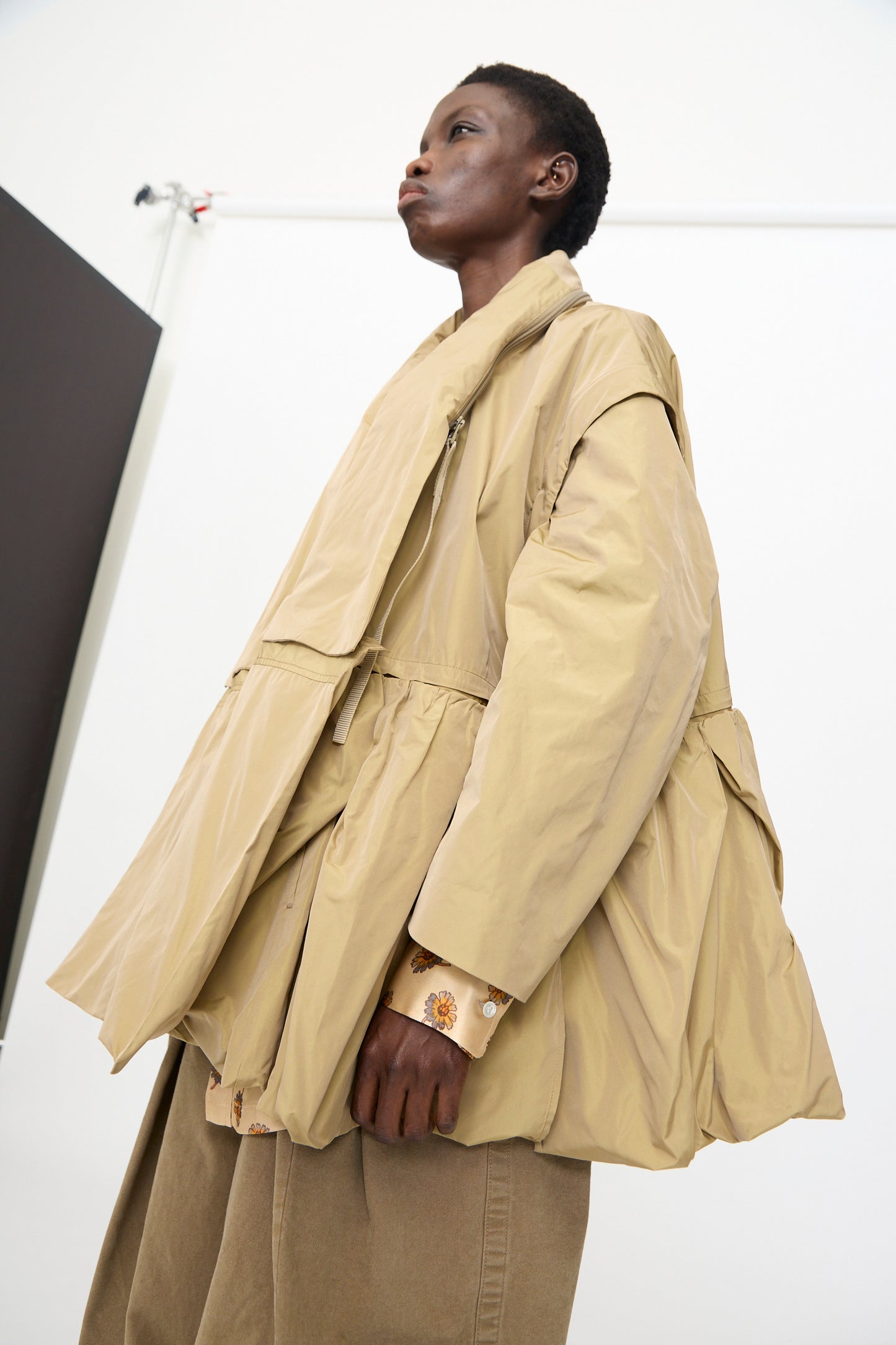 Wearing the TOGA ARCHIVES Memory Taffeta Jacket in Beige, featuring layered details and an adjustable waist, a person pairs it with neutral pants against a plain background.