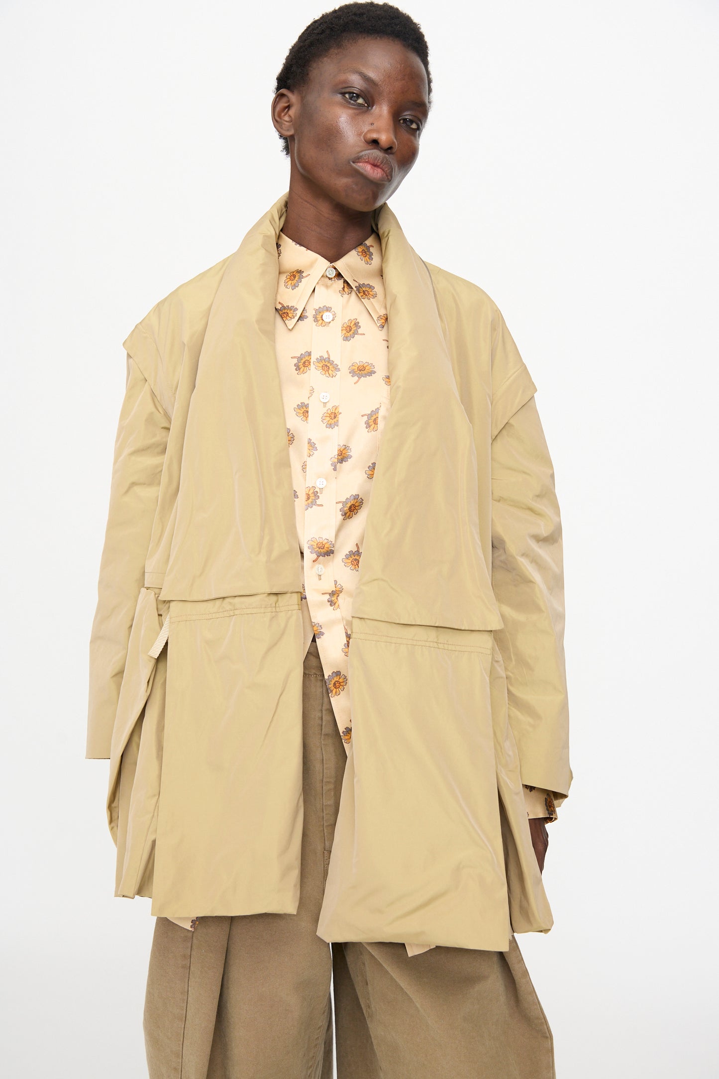 A person wears the TOGA ARCHIVES Memory Taffeta Jacket in Beige, paired with tan wide-leg adjustable waist pants and a light floral-patterned shirt, standing against a plain white background.