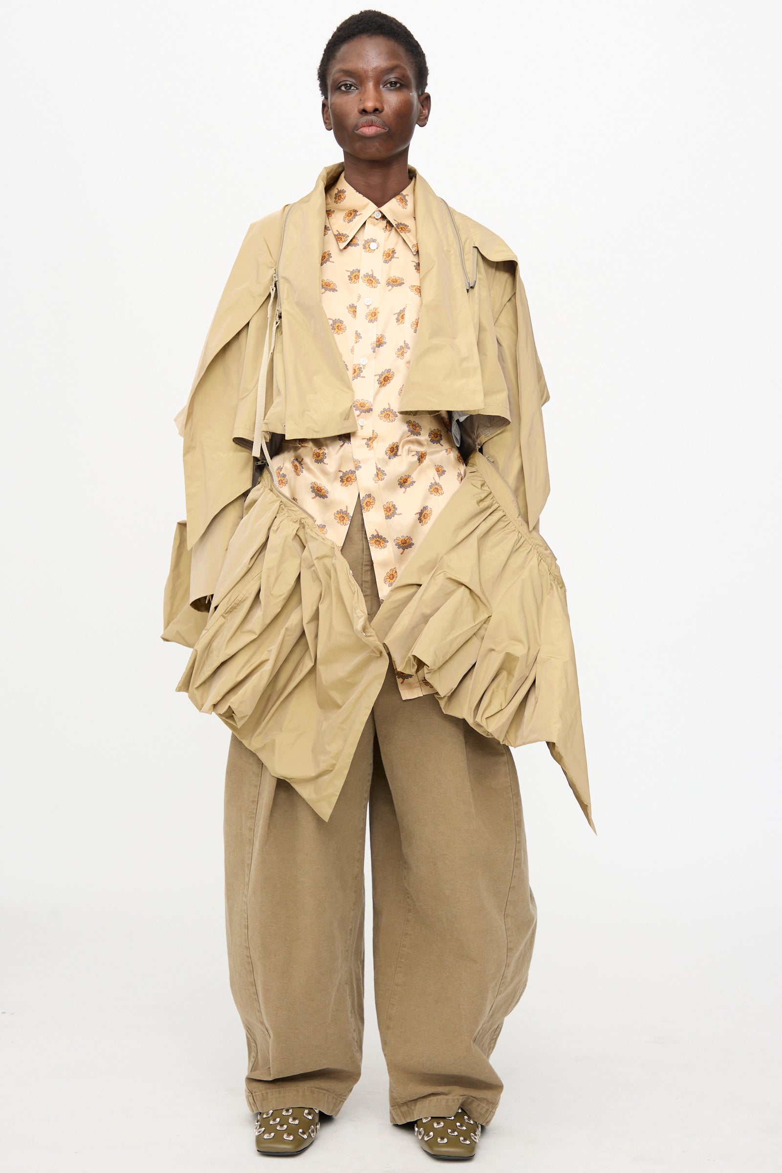 A person stands against a plain background wearing TOGA ARCHIVES' beige Memory Taffeta Jacket, featuring large pockets, paired with a patterned shirt, wide-legged adjustable-waist pants, and studded shoes.