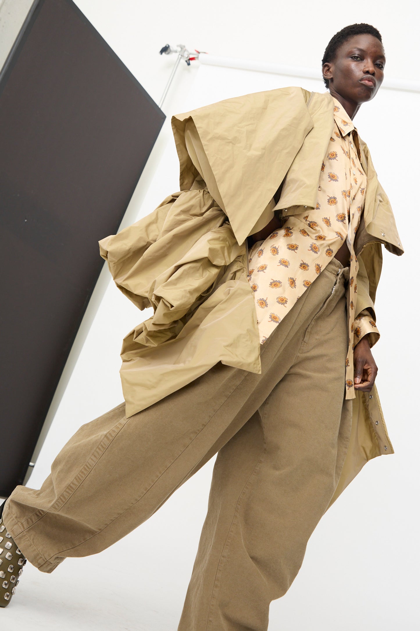 A person wears the TOGA ARCHIVES Memory Taffeta Jacket in Beige, styled with a printed shirt and wide-leg adjustable khaki pants, standing in a studio with a white background and black panel.