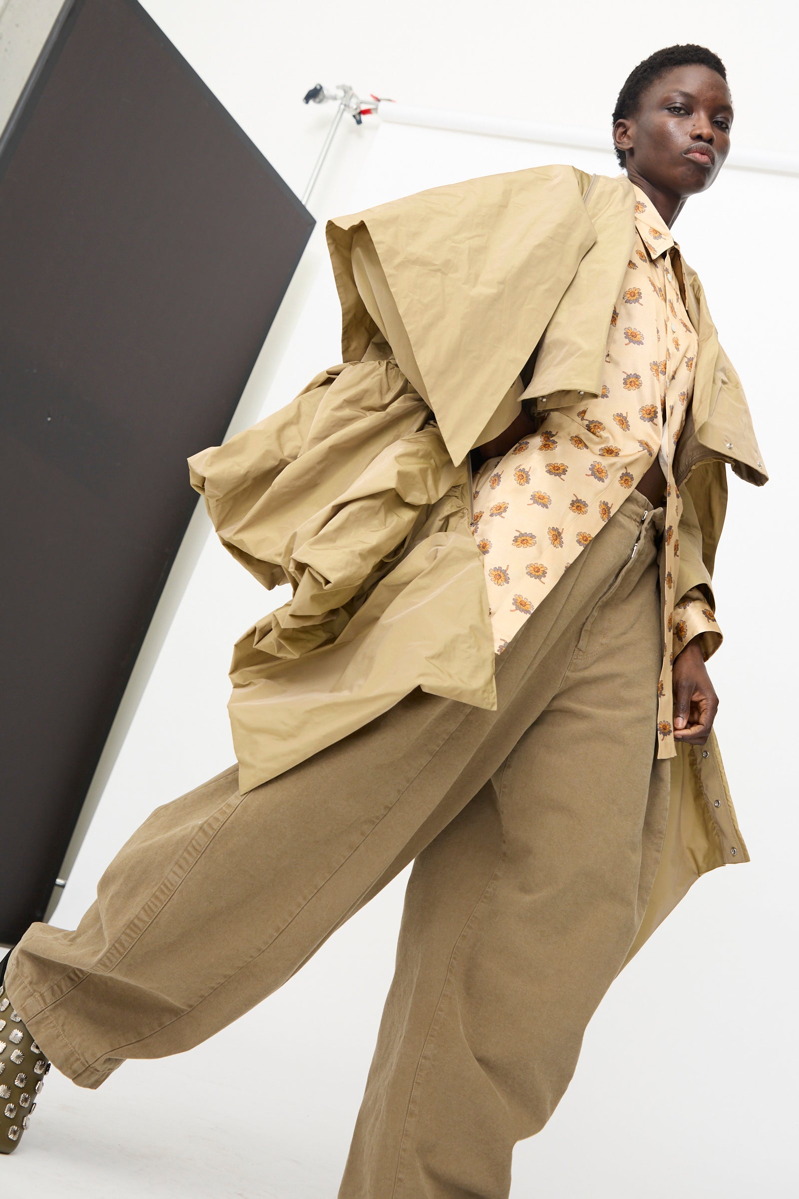 A person wears the TOGA ARCHIVES Memory Taffeta Jacket in Beige, styled with a printed shirt and wide-leg adjustable khaki pants, standing in a studio with a white background and black panel.