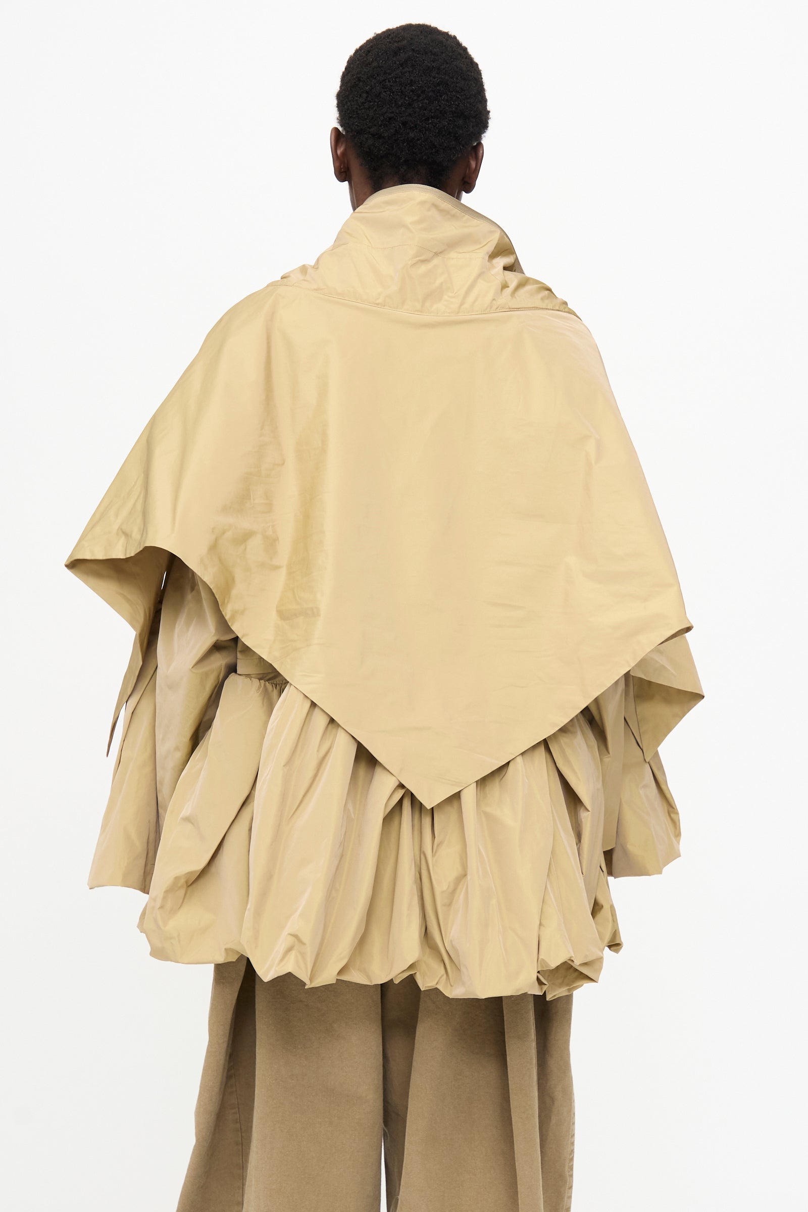 A person faces away from the camera, wearing TOGA ARCHIVES’ beige Memory Taffeta Jacket with layered, gathered fabric and adjustable waist over matching pants, set against a plain white background.