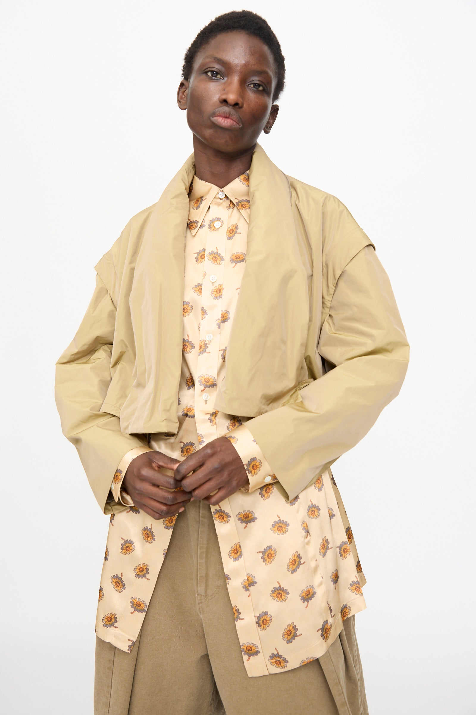 A model wears the TOGA ARCHIVES Memory Taffeta Jacket in Beige over a patterned button-up shirt and tan pants, featuring an adjustable waist, standing against a plain white background.