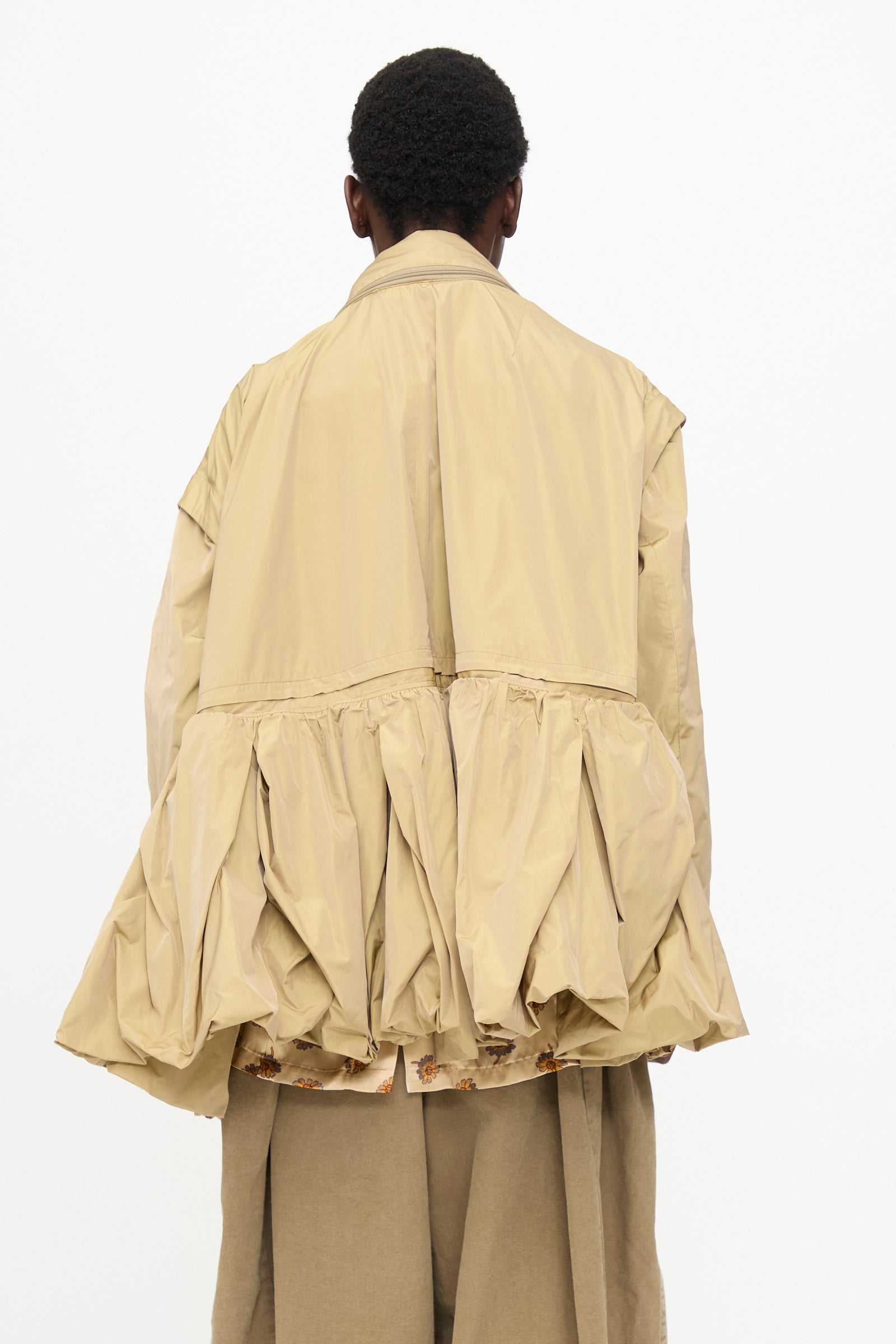 A person stands with their back to the camera, wearing the TOGA ARCHIVES Memory Taffeta Jacket in Beige, featuring a sculptural shape with a gathered, voluminous lower section and adjustable waist, paired with matching wide-legged pants.