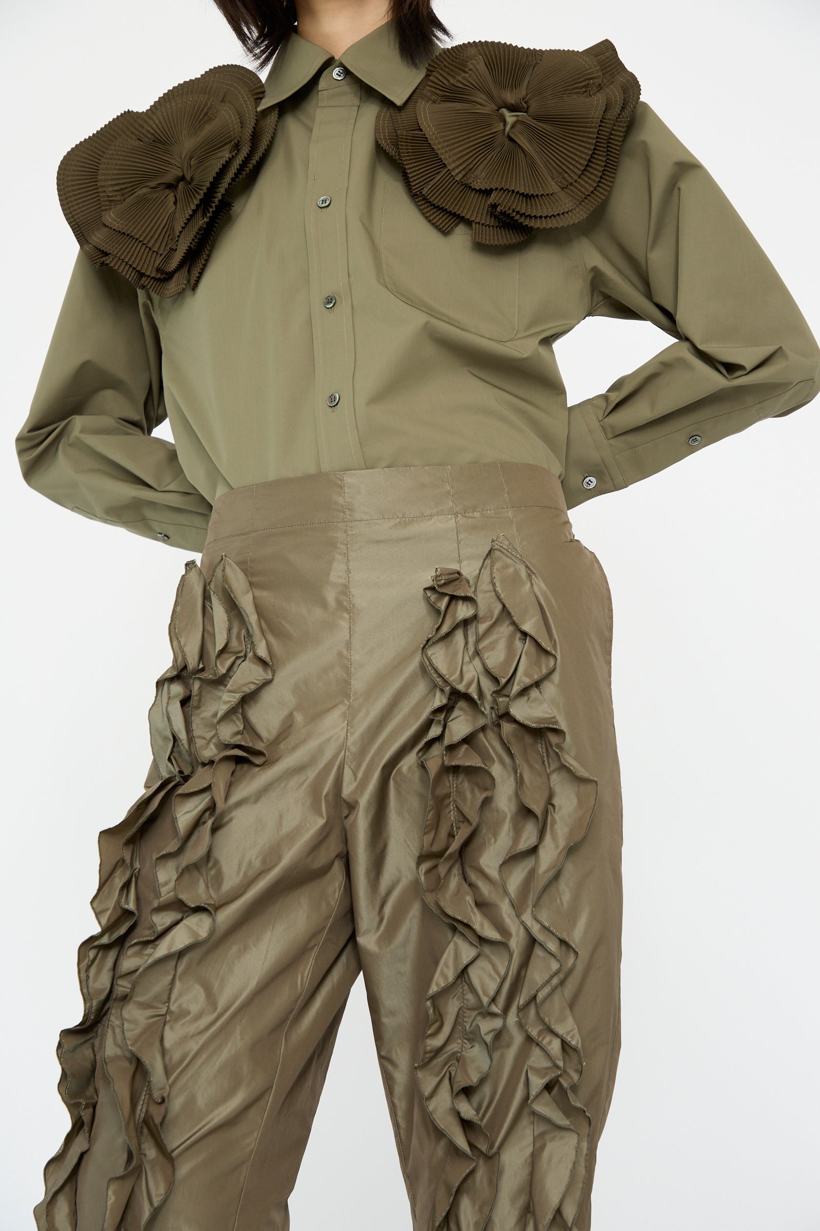 A person in an olive green shirt with large shoulder ruffles paired with TOGA ARCHIVES' Memory Taffeta Pant in khaki, featuring ruffled detailing, against a white background.