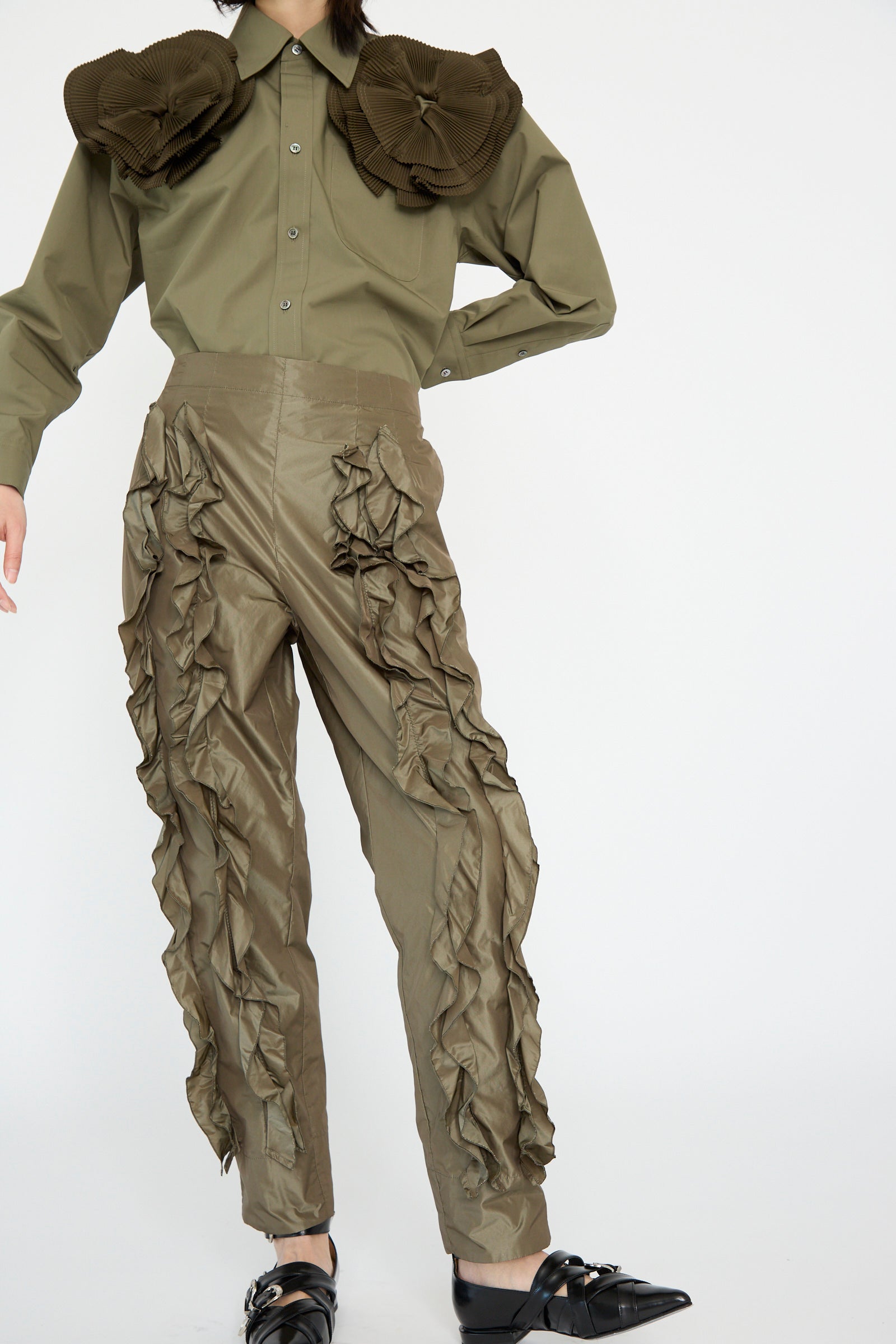 A person in an olive green Memory Taffeta Pant by TOGA ARCHIVES wearing a shirt with large ruffles on the shoulders and sides, against a white background. 