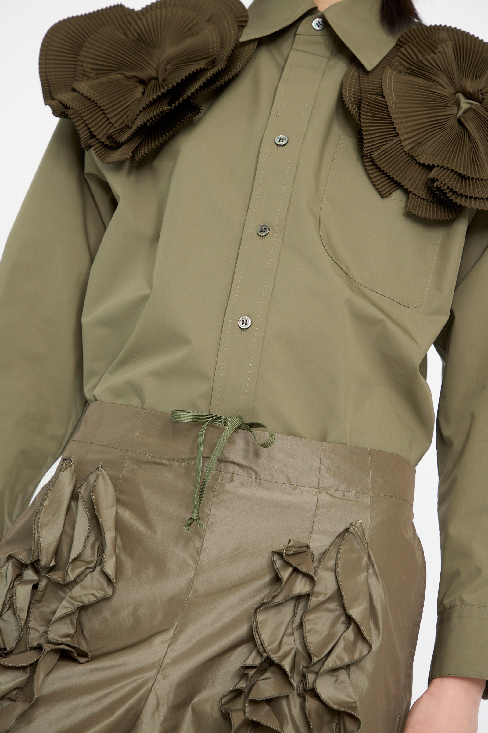 Someone wearing a green button-up shirt with large fabric embellishments on the shoulders paired with TOGA ARCHIVES' Memory Taffeta Pant in Khaki, which features taffeta ruffled detailing.