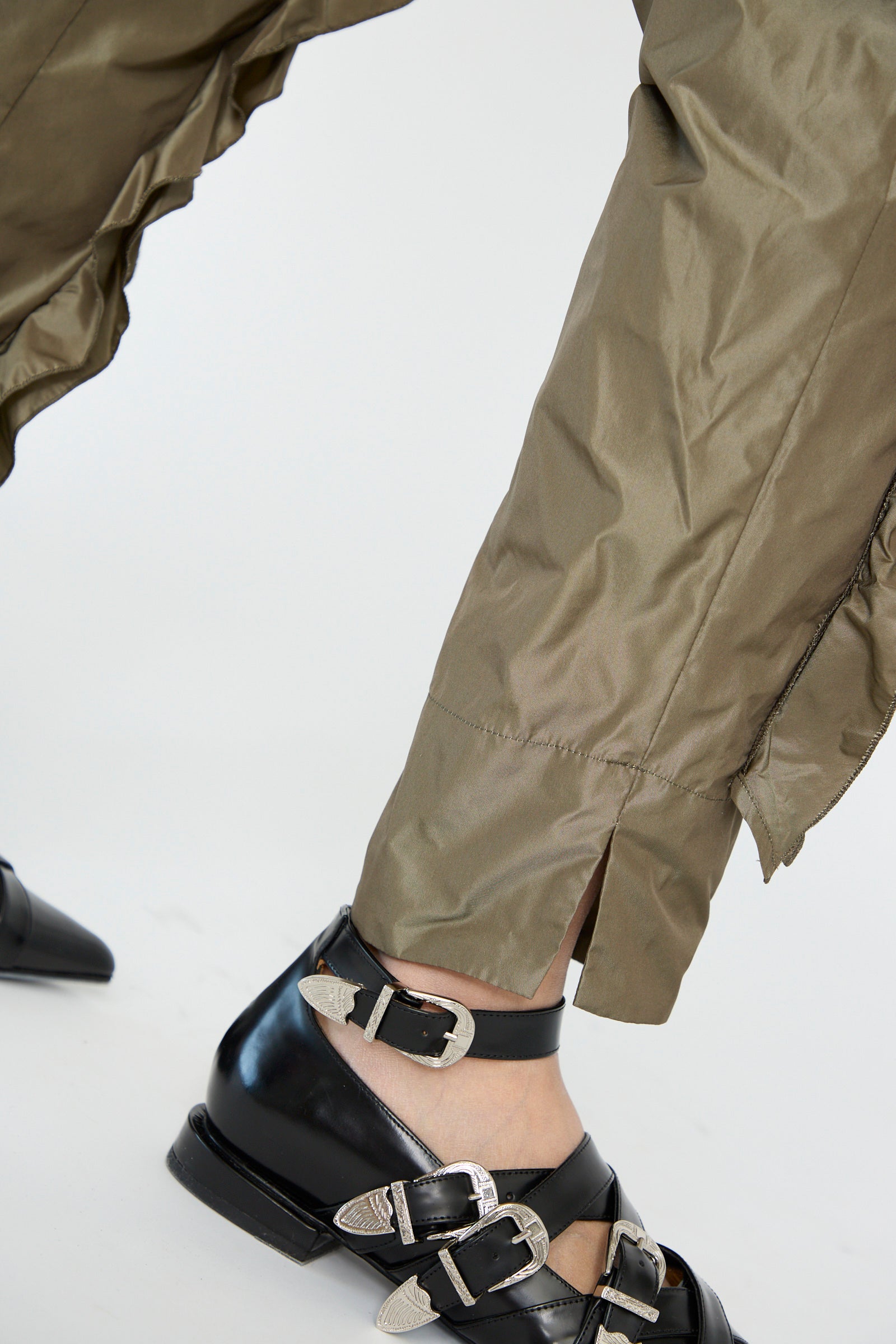 Close-up of someone wearing the TOGA ARCHIVES Memory Taffeta Pant in Khaki, paired with black shoes with silver buckles against w white background.