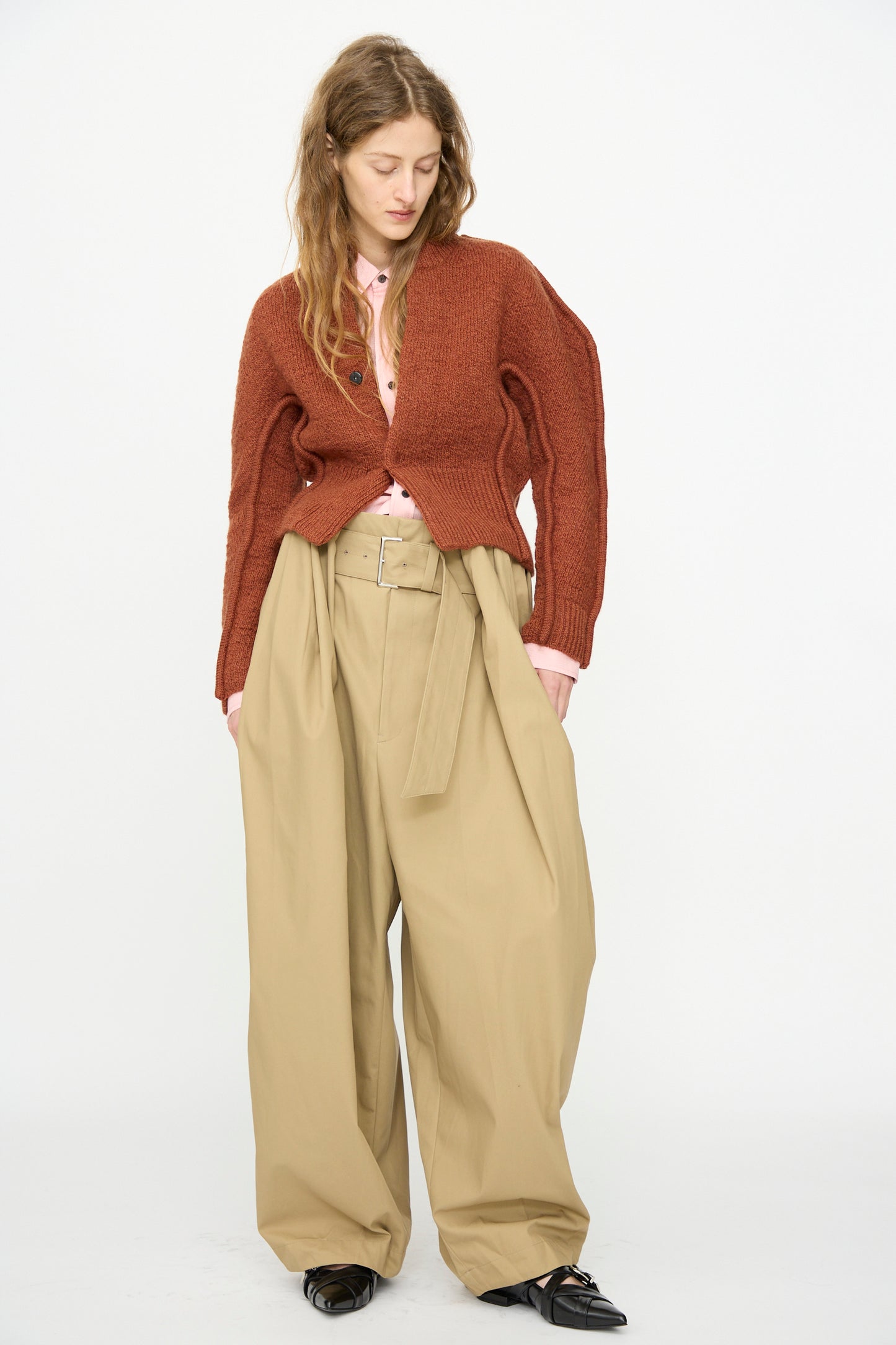 A person wears the TOGA ARCHIVES Motif Knit Cardigan in Brown over a pink shirt, paired with wide beige belted trousers and black shoes, standing against a white background. A sparkling flower brooch accents the structured look.
