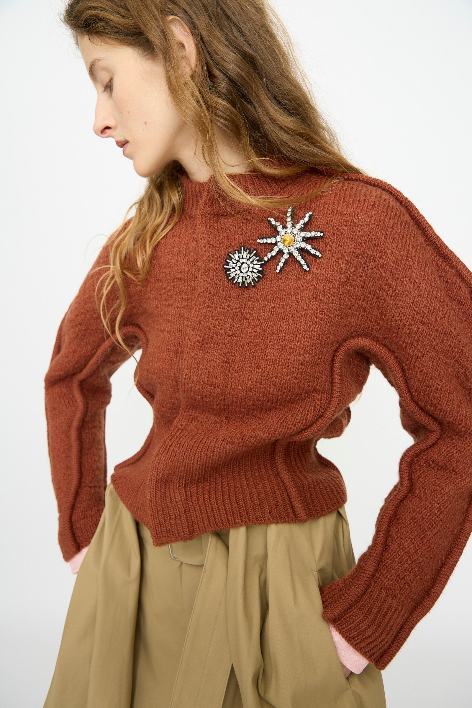 Woman wearing the TOGA ARCHIVES Motif Knit Cardigan in Brown, styled with beige high-waisted pants, featuring bejeweled flower brooch accents, against a plain light background.