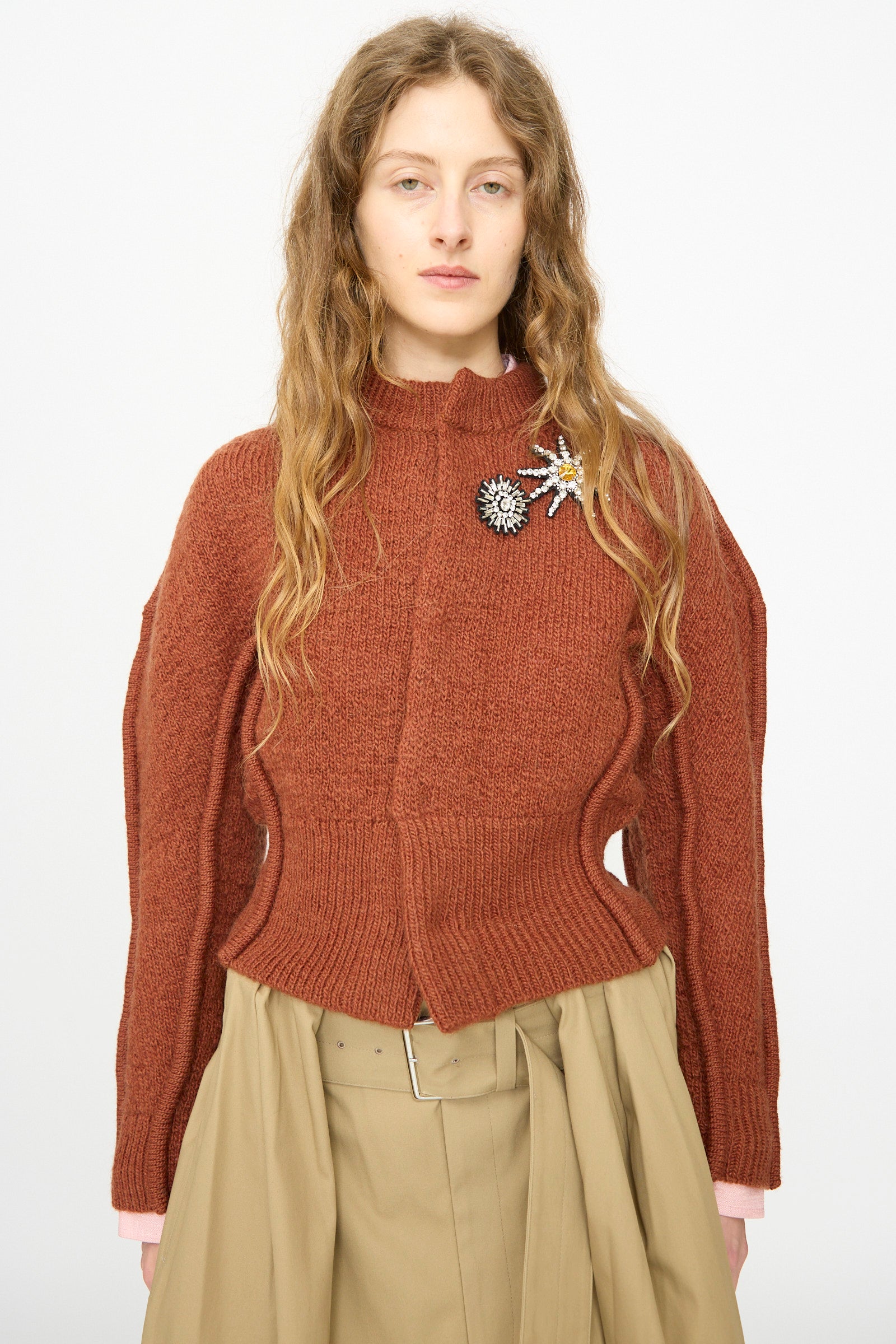 A person with long wavy hair wears the TOGA ARCHIVES Motif Knit Cardigan in Brown, featuring embellished flower brooches, paired with a beige skirt and wide belt, standing against a plain light background.