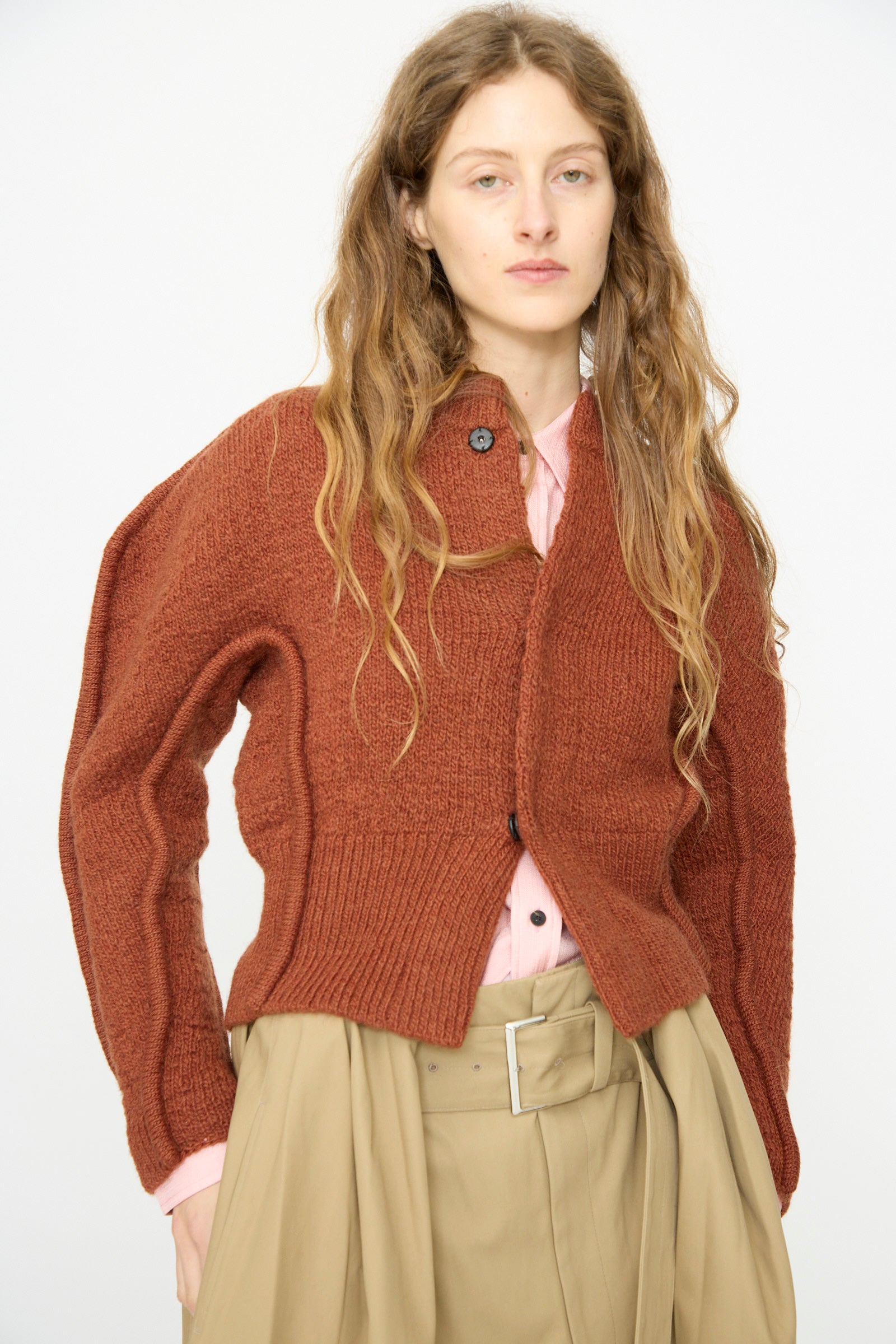 A person with long wavy hair wears the TOGA ARCHIVES Motif Knit Cardigan in Brown over a pink shirt and beige high-waisted pants, standing against a plain white background.