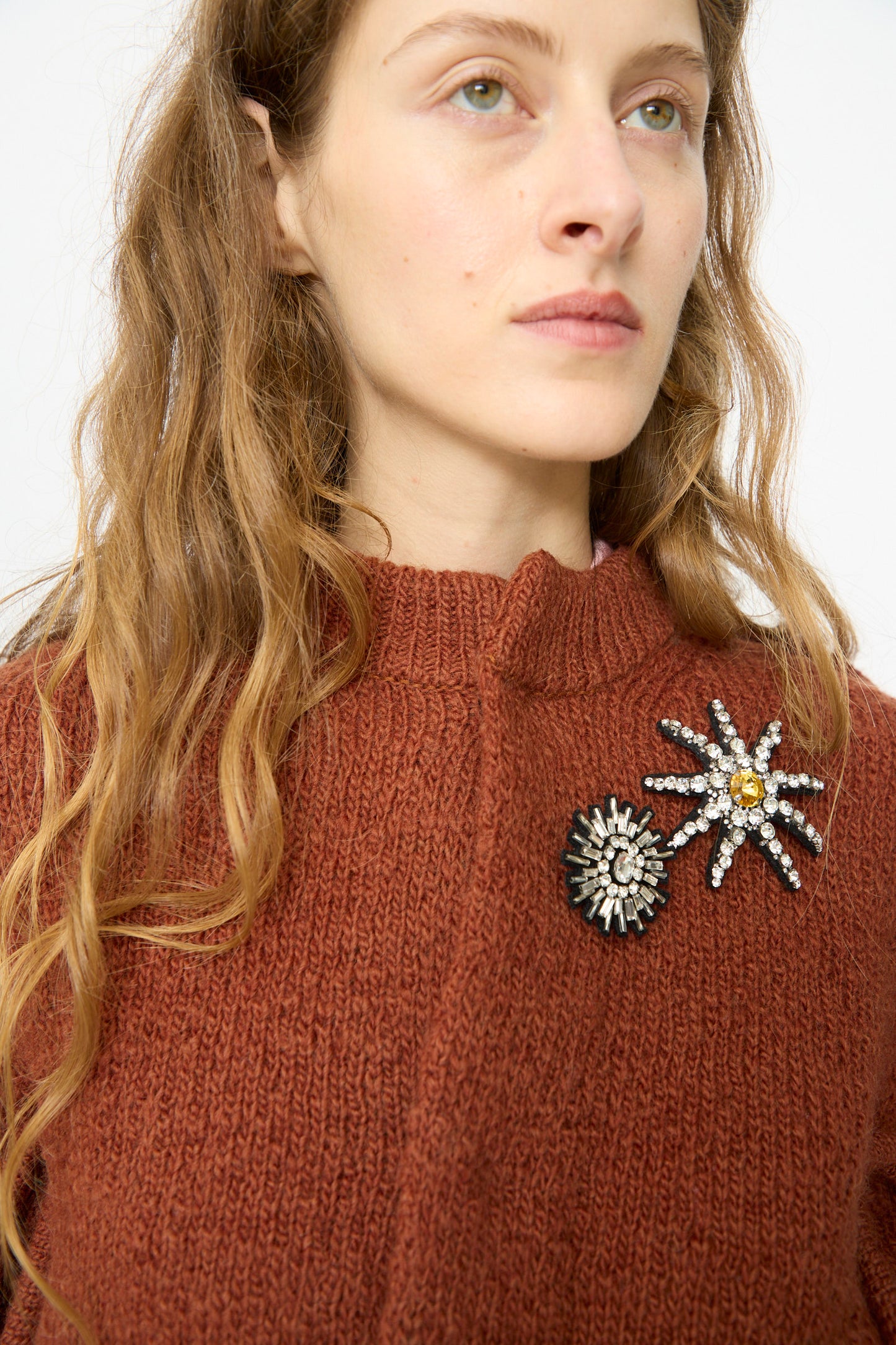 A woman wears the TOGA ARCHIVES Motif Knit Cardigan in Brown, a wool long sleeve piece with a structured fit, featuring two brooches—a round one and a jeweled flower—pinned to the left chest.