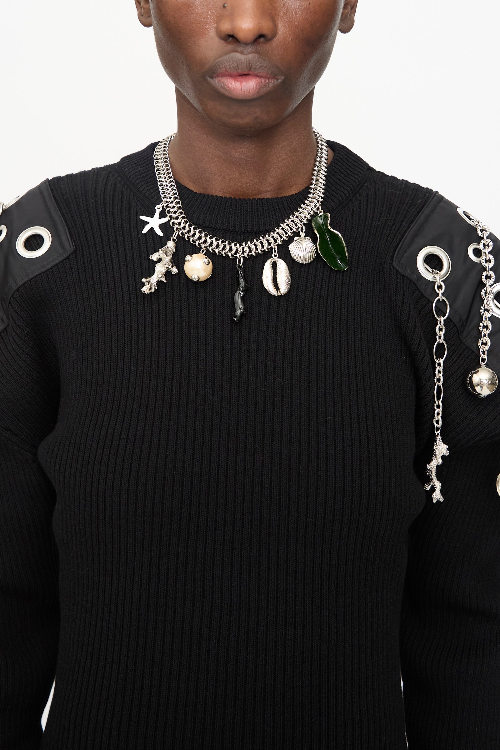 A person wears a black ribbed sweater with chain details and the TOGA ARCHIVES Motif Necklace in Silver, featuring a toggle clasp on a silver chain adorned with shells, a leaf, and sea-themed charms.