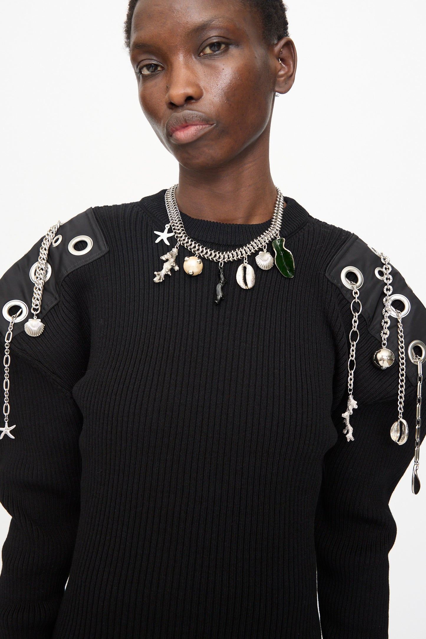 Person wearing a black ribbed sweater with metallic and shell-embellished shoulders and the Motif Necklace in Silver by TOGA ARCHIVES, standing against a plain white background.
