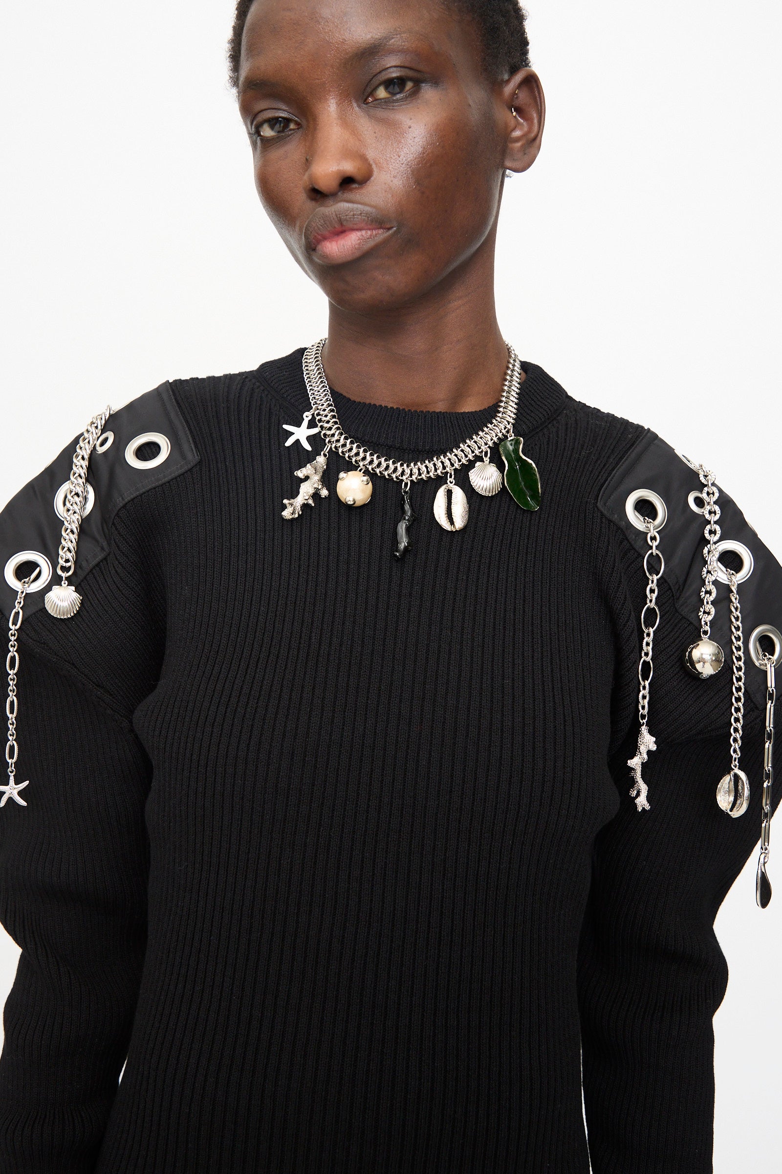 Person wearing a black ribbed sweater with metallic and shell-embellished shoulders and the Motif Necklace in Silver by TOGA ARCHIVES, standing against a plain white background.