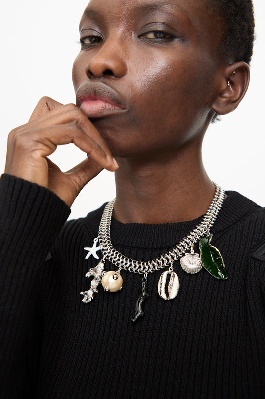 A person wears the Motif Necklace in Silver by TOGA ARCHIVES, featuring a chunky silver chain with eclectic charms and a toggle clasp, styled over a black ribbed sweater.