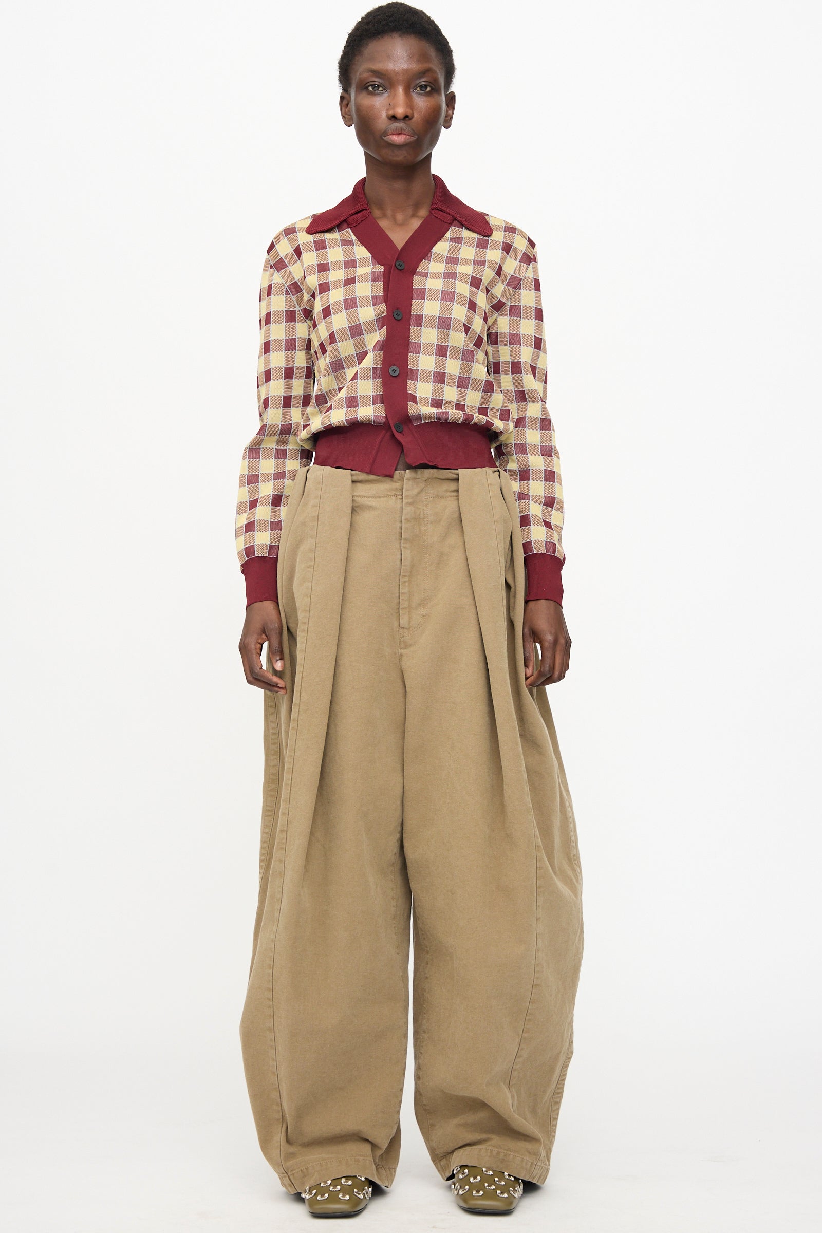 A person stands against a white background wearing the TOGA ARCHIVES Oxford Wide Pants in Beige with a burgundy and beige plaid shirt featuring large buttons, completed with studded shoes.